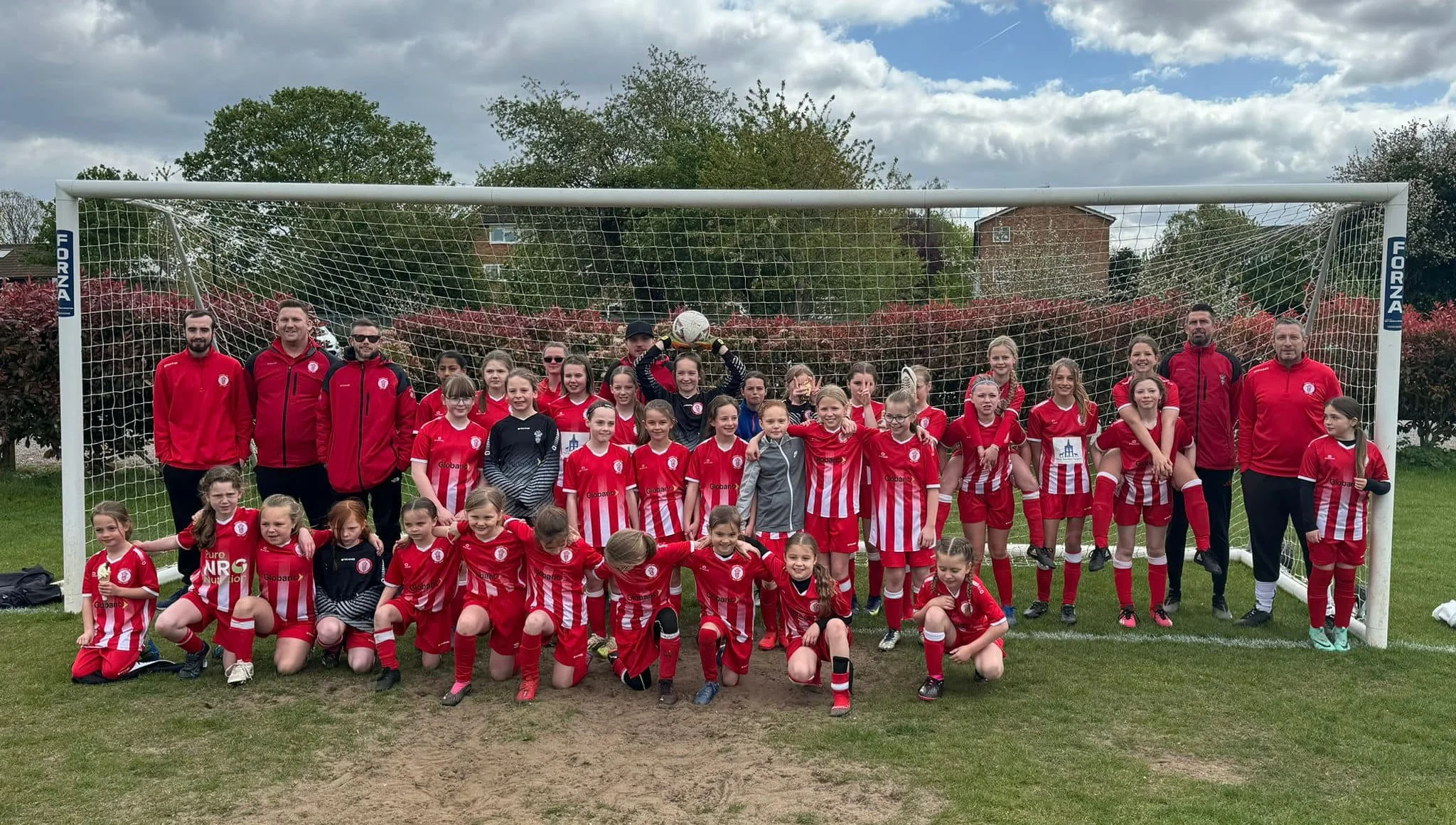 Group of young female football players and their coaches posing in front of a goal after a game, with players wearing red and white striped kits and coaches in red jackets, outdoors on a cloudy day.