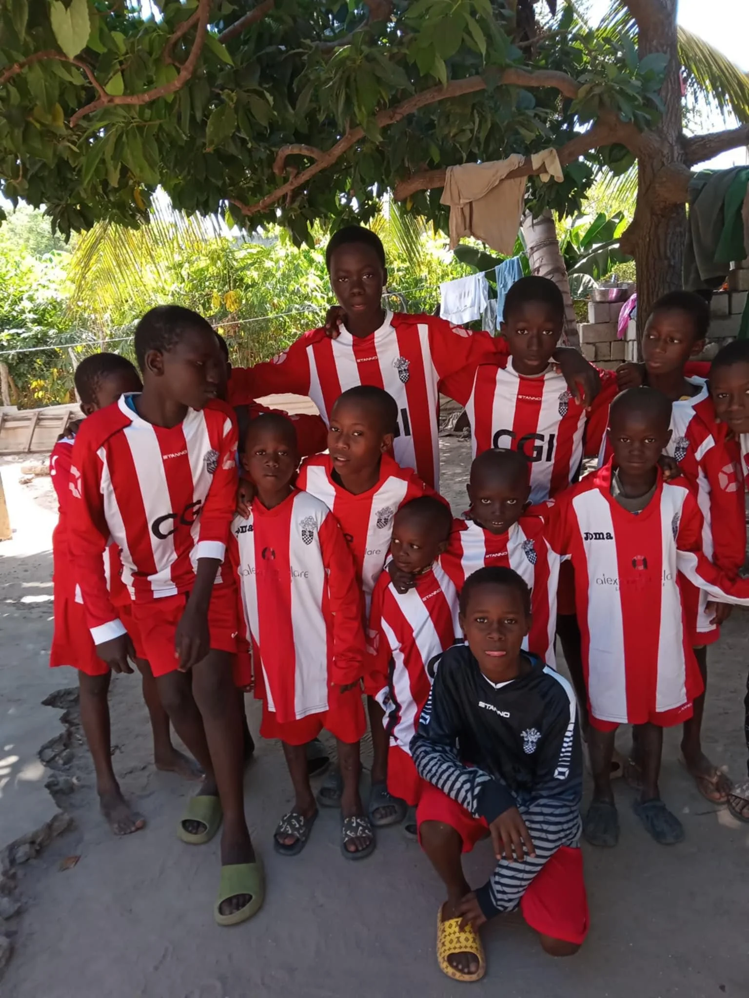 Group of young African children and a teenager in red and white soccer jerseys posing under a tree outdoors, with some children wearing sandals and slippers, and clothes hanging on a line in the background.