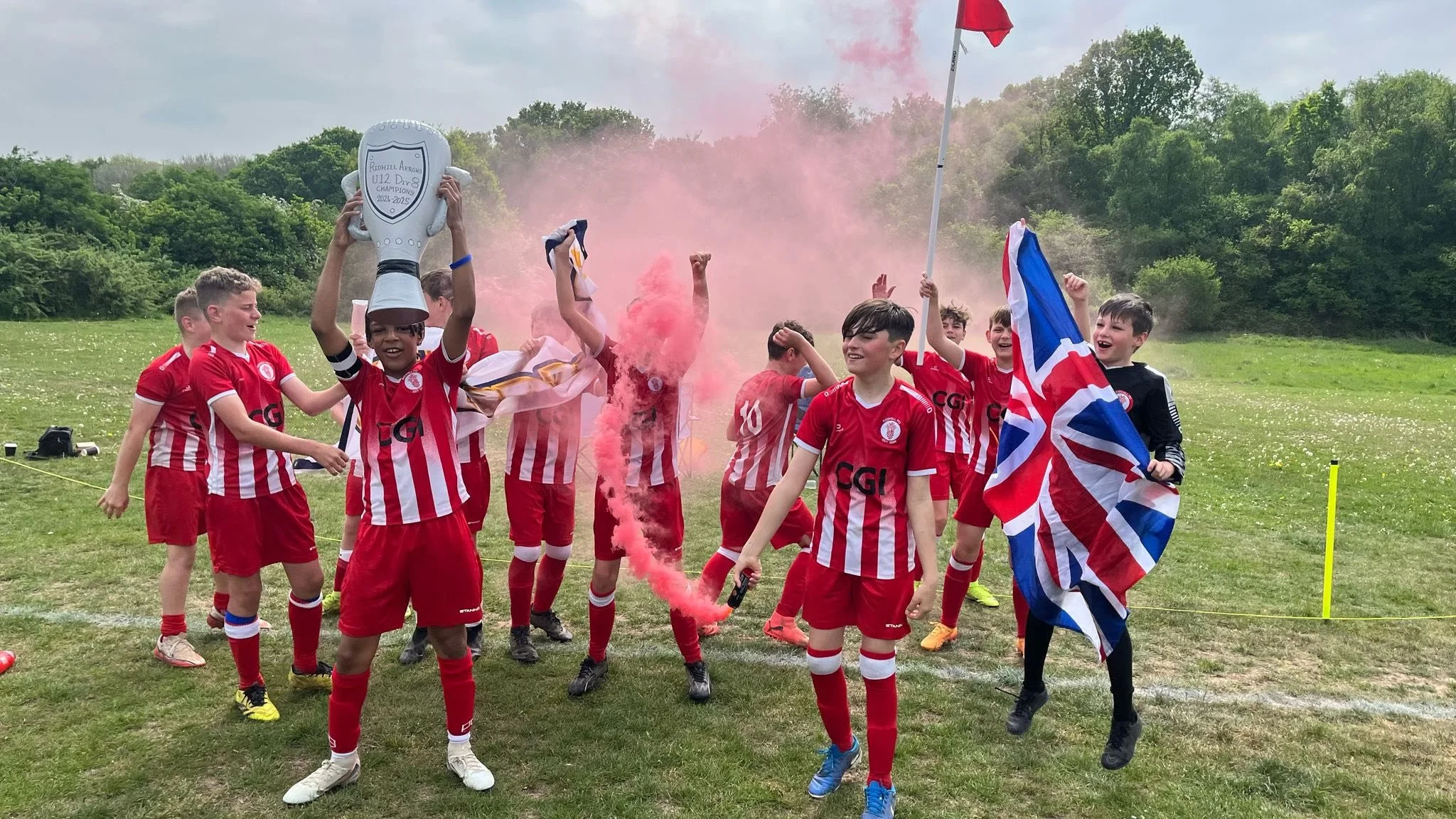 Young soccer players celebrating on a field, one holding a trophy and others holding flags and smoke flares, wearing red and white striped uniforms.