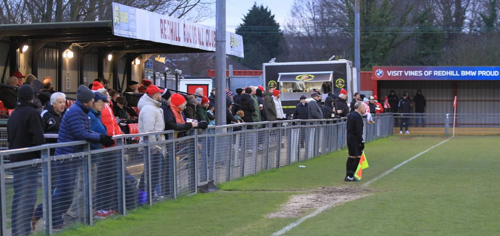Spectators watching a soccer game from the stands at a field, some wearing winter clothing and red Santa hats, with a referee standing near the sideline.