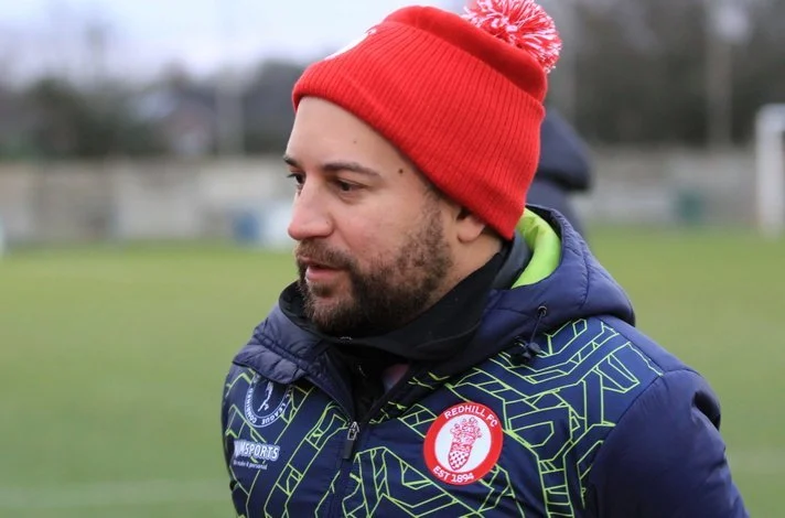 A man wearing a red knit beanie with white pom-pom, a blue jacket with a sports logo, standing outdoors on a grassy field.