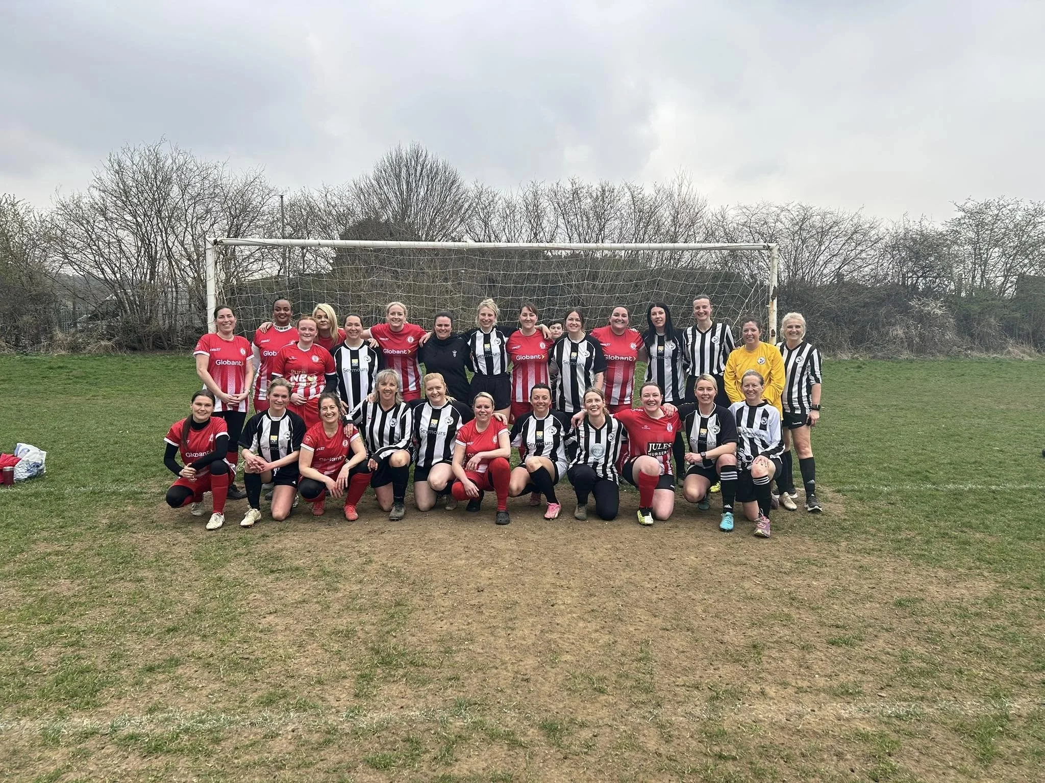 Group of women soccer players posing on a field, some wearing red jerseys and others in black and white stripes, with a goalpost behind them.