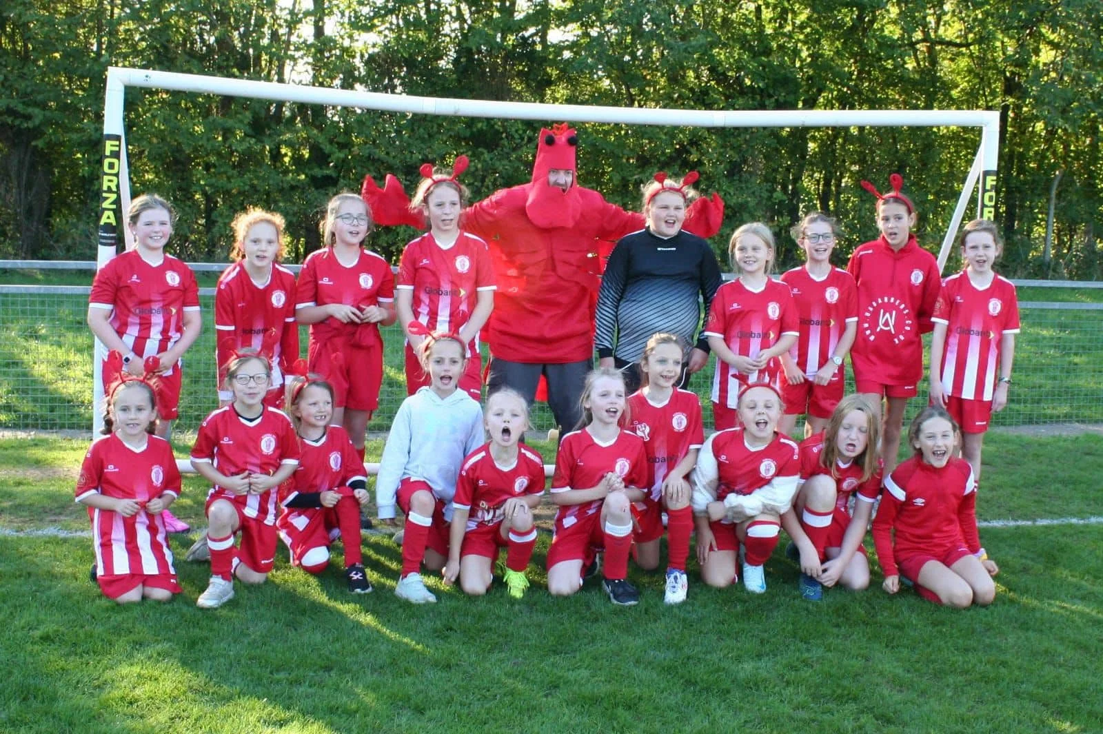 A group of young girls in red soccer uniforms and a person in a lobster costume posing for a photo on a soccer field.