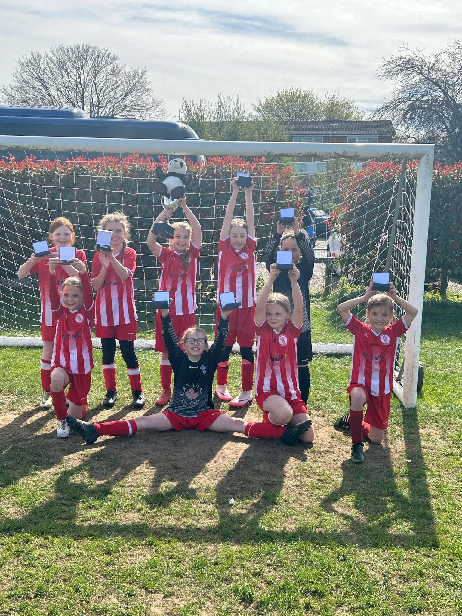 Girls' youth soccer team celebrating with their medals and trophy on a soccer field, standing in front of a goalpost on a sunny day.