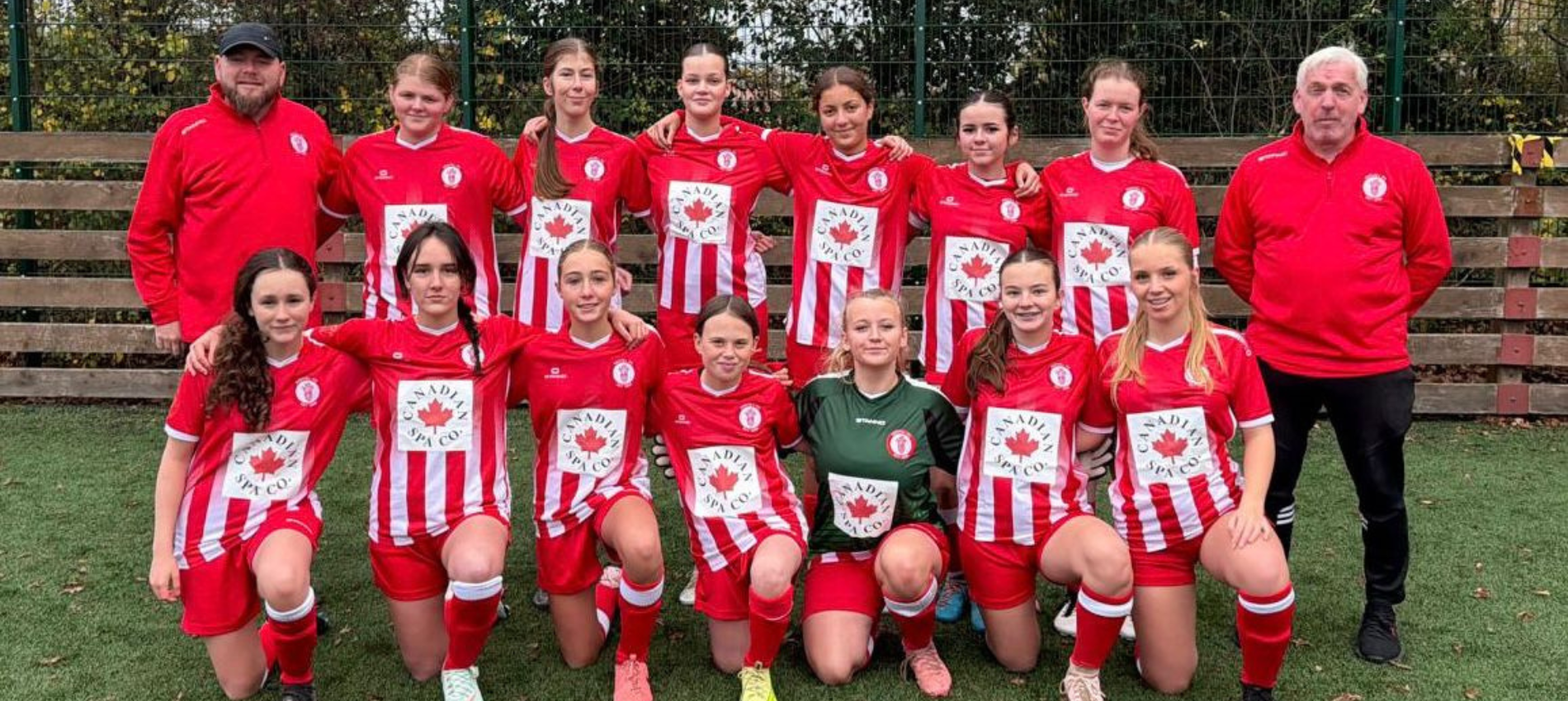 A girls' soccer team in red and white striped uniforms, along with two coaches, posing on a soccer field.