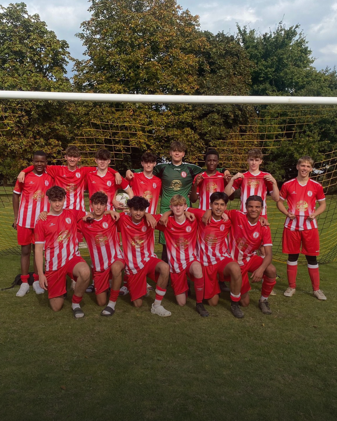 A youth football team in red and white kits, with one player in a green goalkeeper jersey, standing and kneeling in front of a goal on a grassy field with trees in the background.