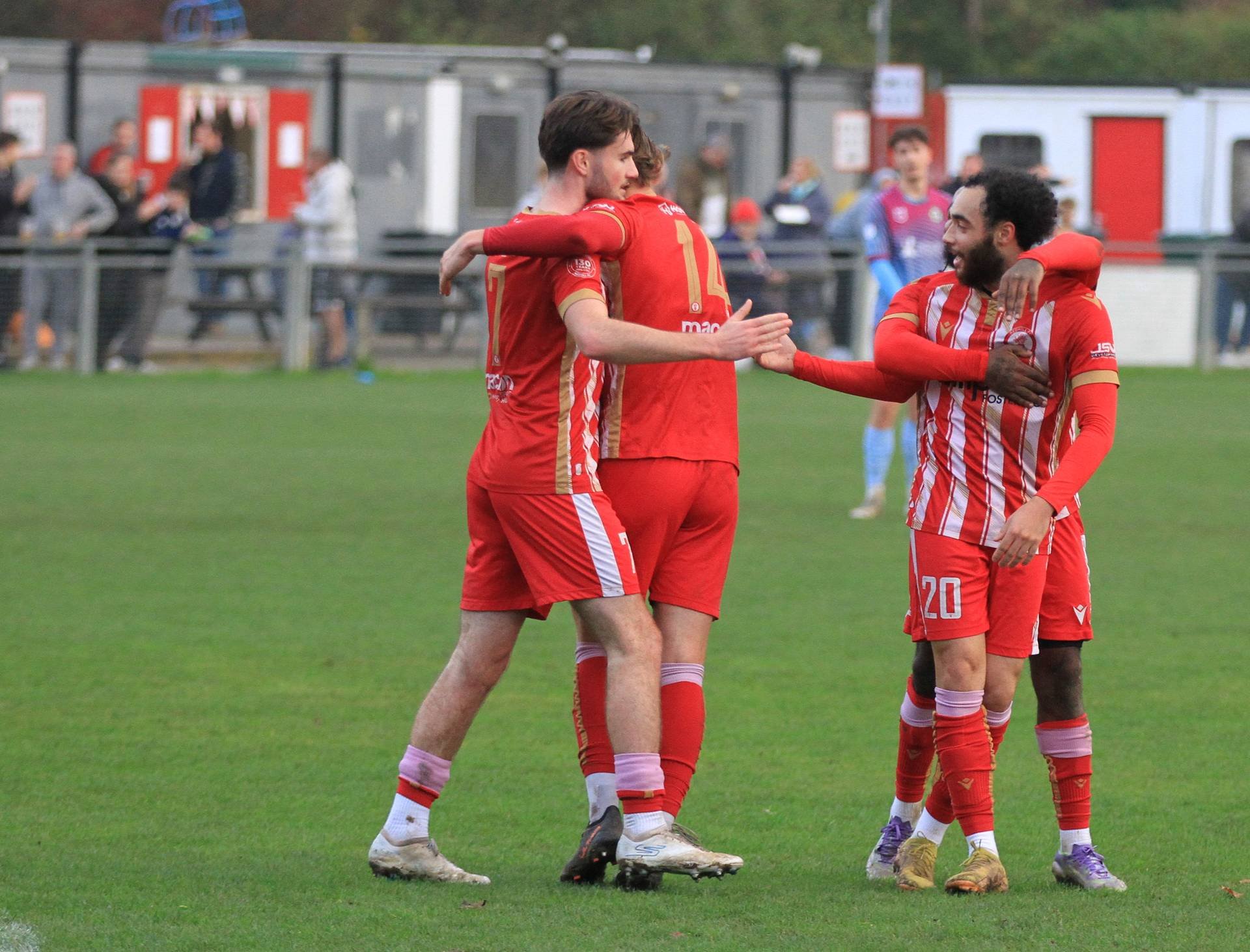 Four soccer players in red uniforms celebrating on the field, embracing and smiling.