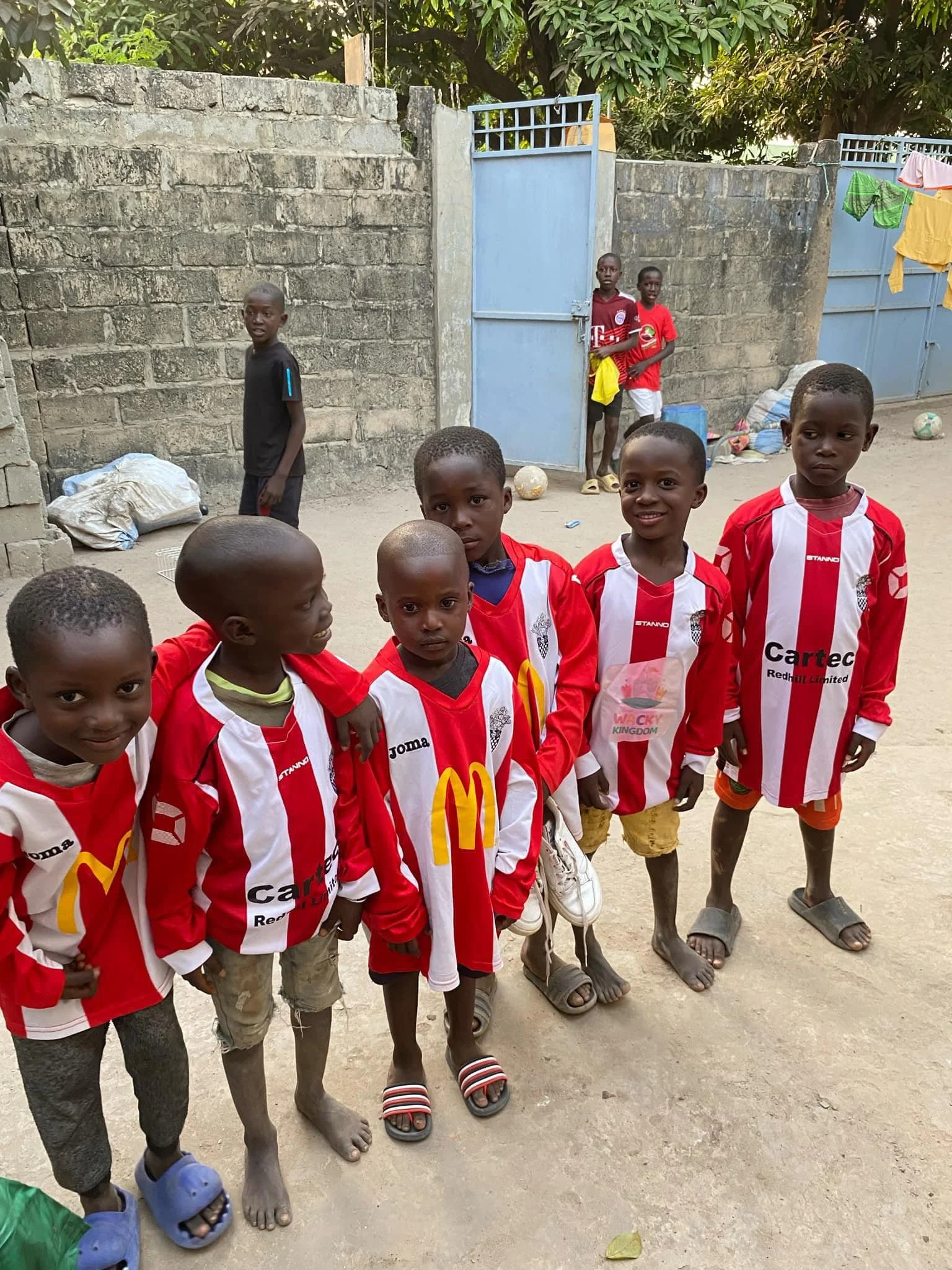 Seven young boys wearing red and white soccer jerseys standing together on a dusty street, with some barefoot or wearing sandals, and a brick wall with a blue gate in the background.