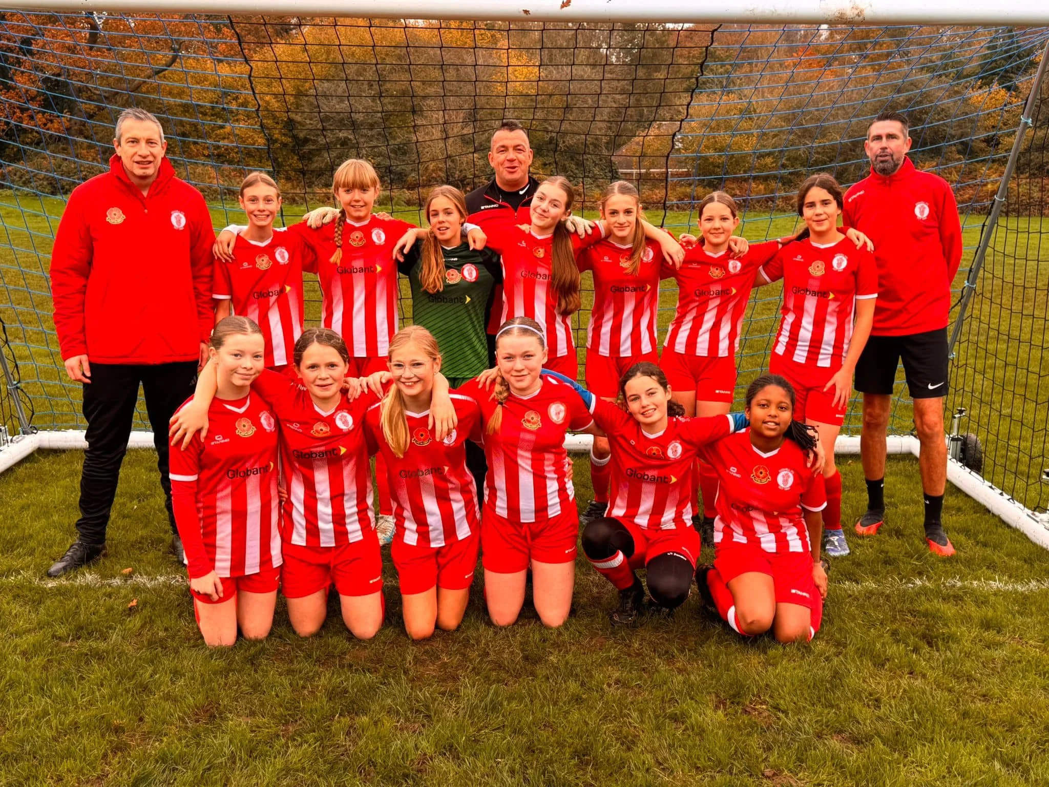 A girls' soccer team photo with players and coaches posing in front of a goal net outdoors during fall with trees in the background, all wearing red and white striped uniforms.