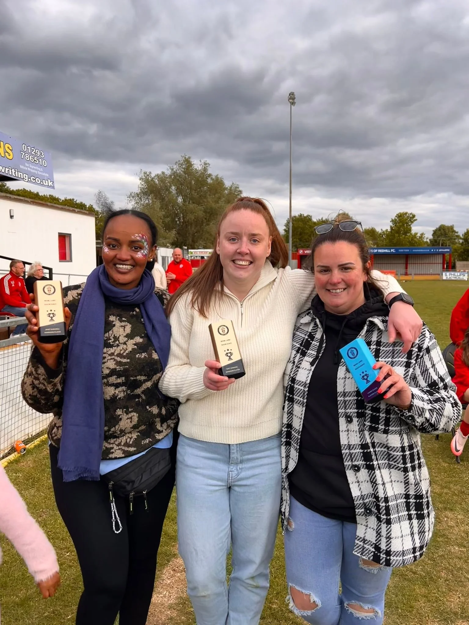 Three women standing together outdoors at a sports field, smiling, holding awards. Two women are holding black awards, and one woman is holding a blue award, with cloudy skies above.