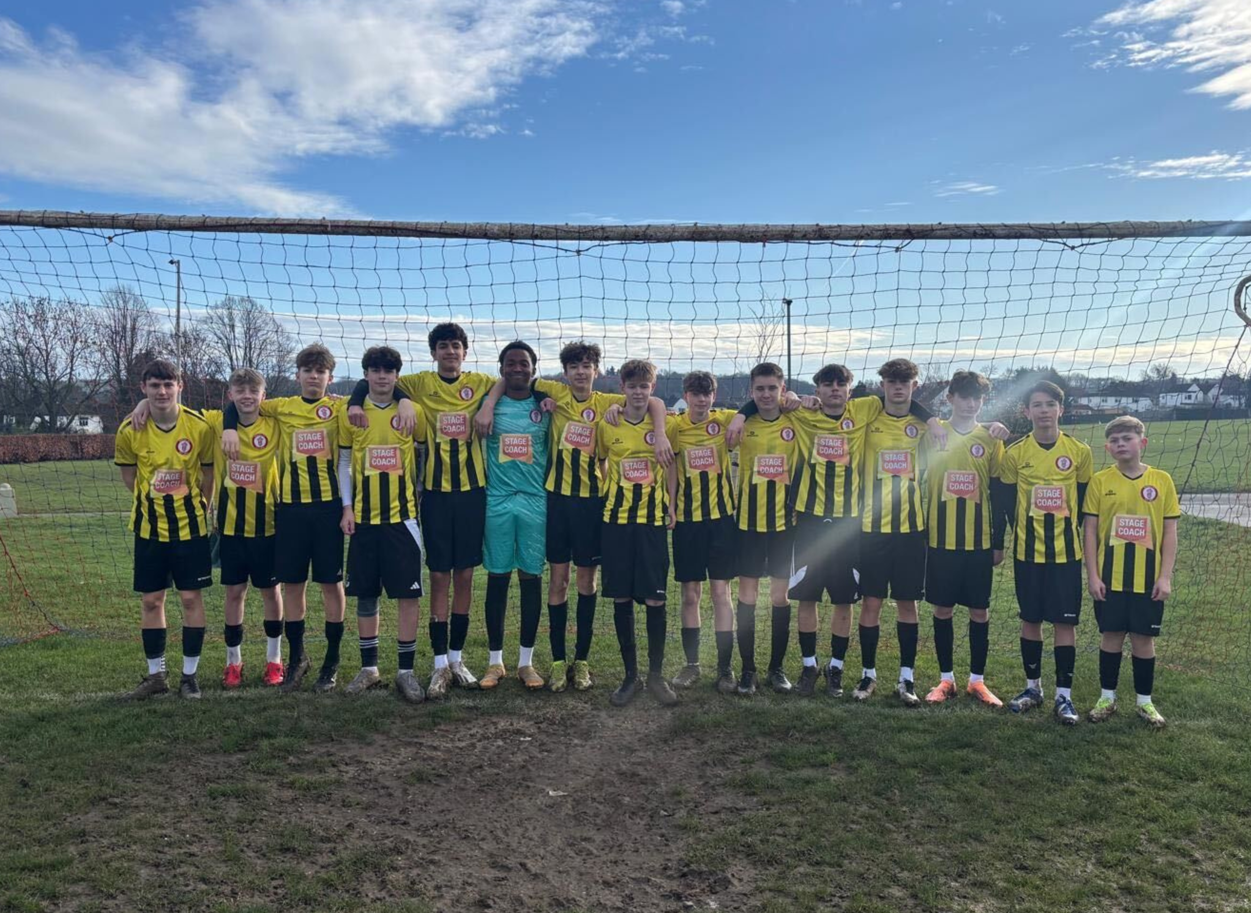 A youth football team of 14 players and one coach or goalkeeper standing in front of a goalpost on a football pitch. They are wearing yellow and black striped jerseys and black shorts. The sky is partly cloudy.