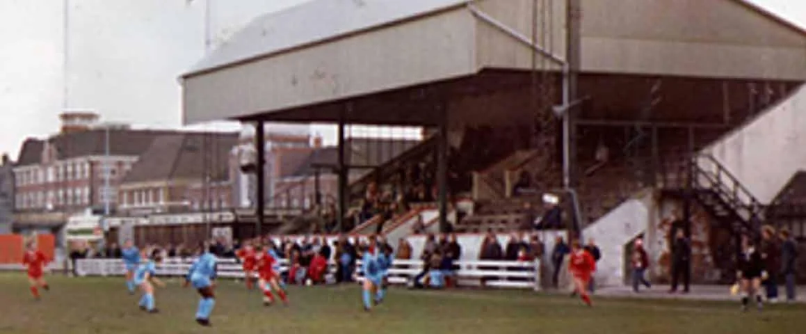 Photograph of a soccer match with players in blue and red jerseys on a grassy field, spectators sitting and standing near a grandstand, and neighboring buildings in the background.