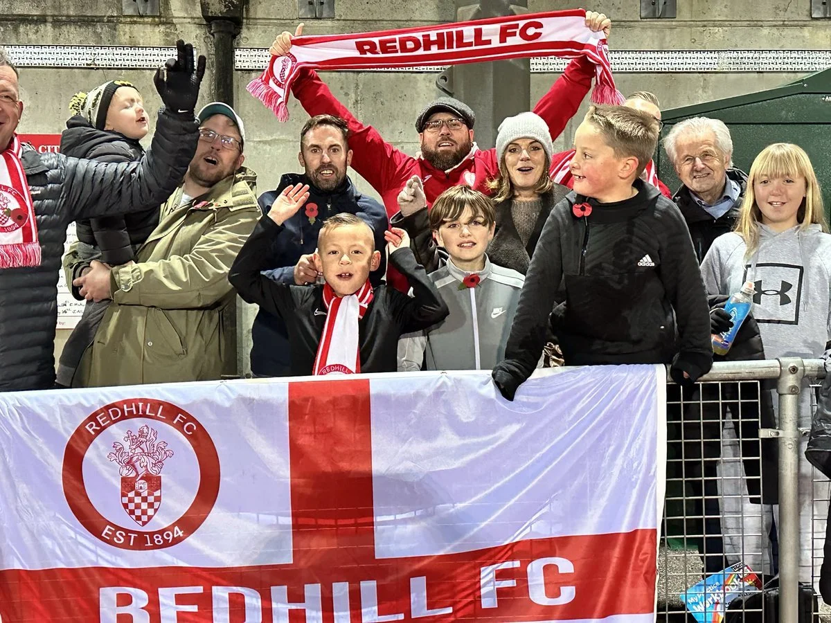 Group of people, including children and adults, standing behind a Redhill FC banner, some holding red and white scarves, at a match.