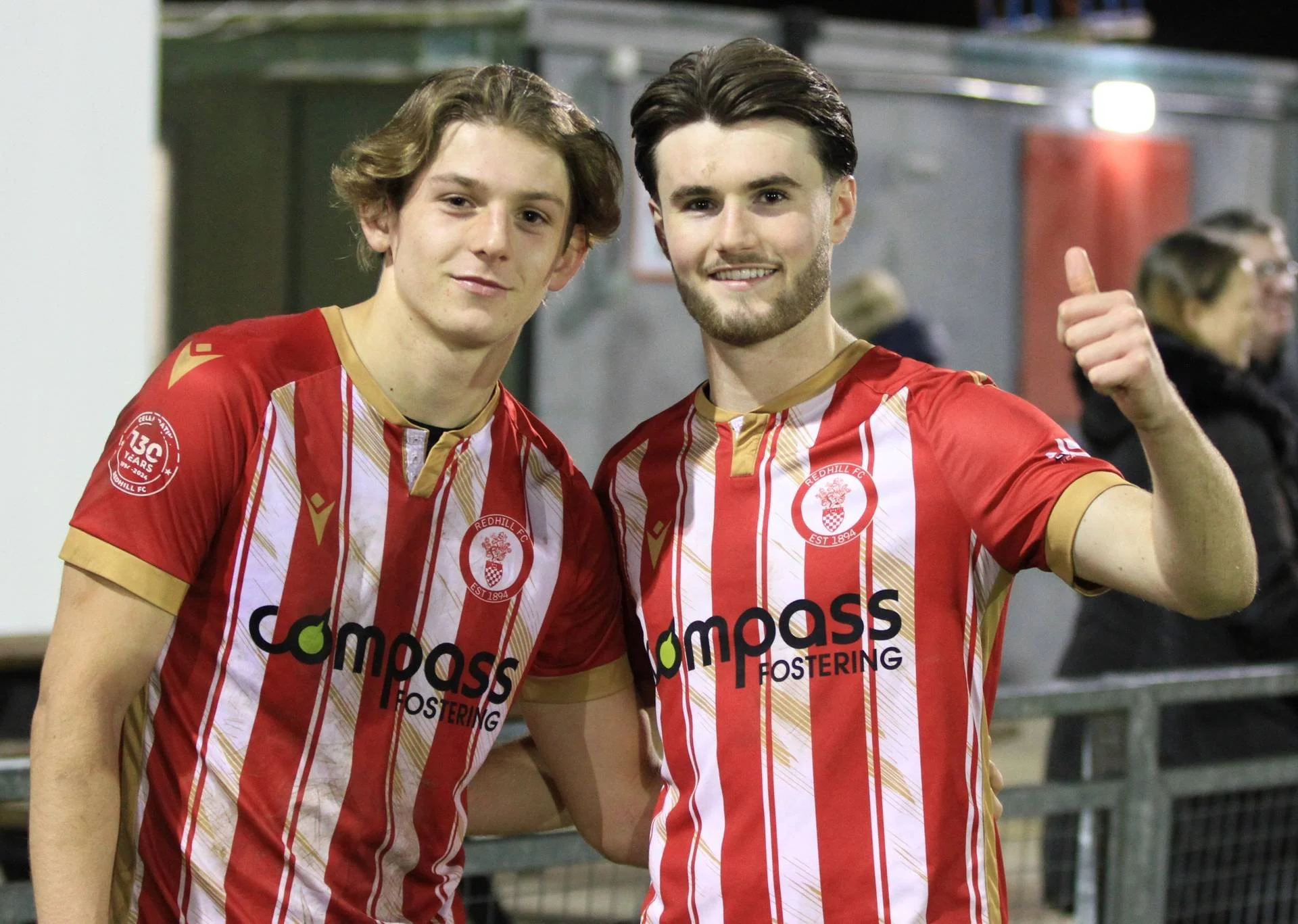 Two young men in red and white striped football kits are smiling and posing for a photo, one making a thumbs-up gesture.