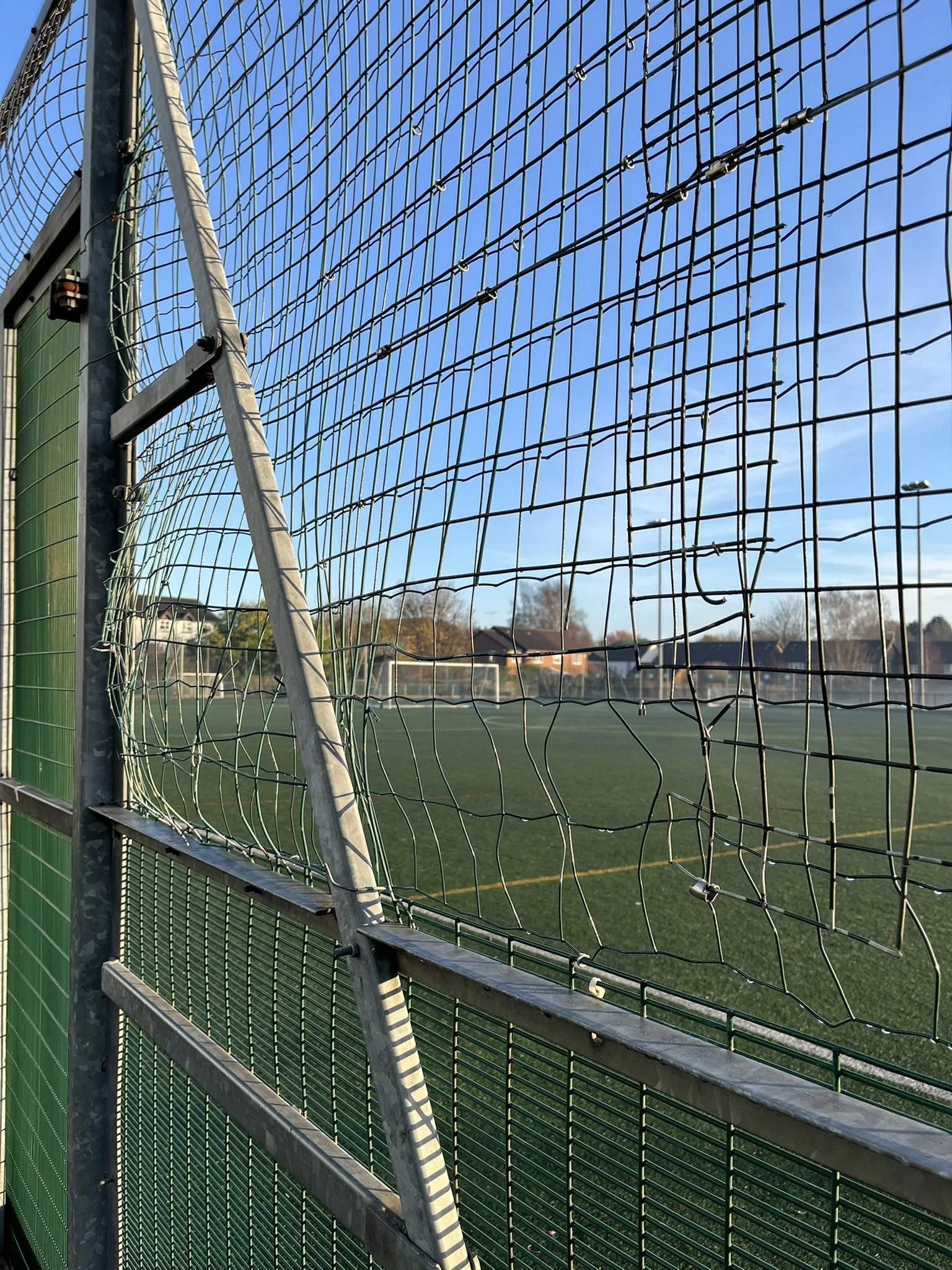 Close-up of a damaged chain-link fence at a sports field with a goal in the background and a clear blue sky.
