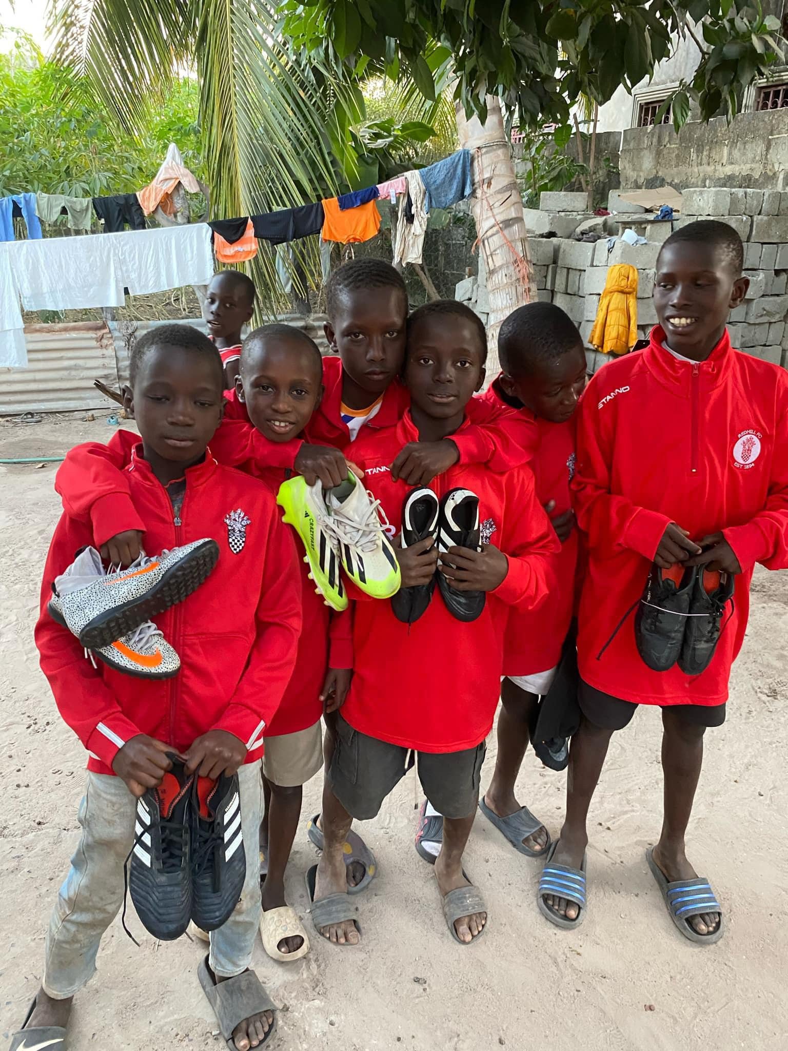 Group of six young boys wearing red jackets, holding sports shoes, standing on a sandy ground in an outdoor setting with trees, laundry hanging, and a wall in the background.