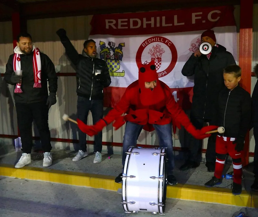 A group of football fans, some wearing Redhill F.C. gear, gather at a game or event behind a Redhill F.C. banner. One person dressed in a Lobster costume plays a drum, while another holds a megaphone. Everyone appears to be celebrating or cheering.