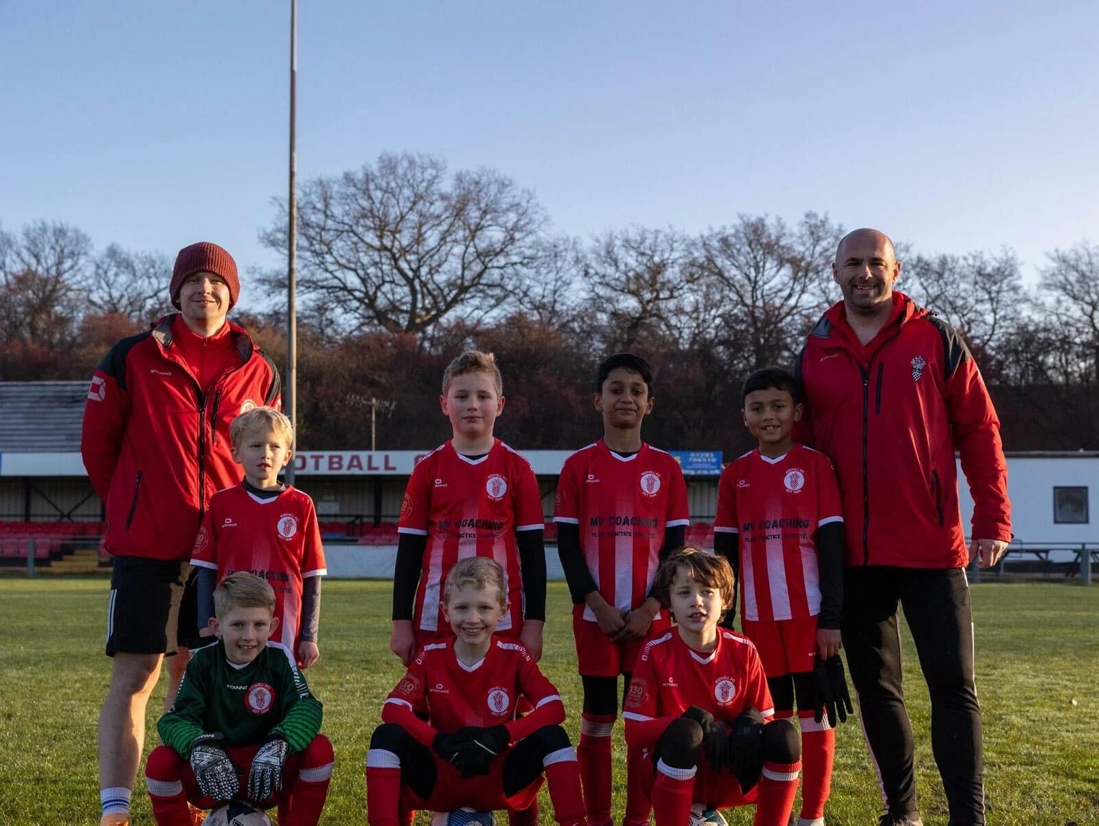A youth football team with two coaches on a pitch, posing for a group photo. The team members are wearing red and white kit, with one child in a green goalkeeper kit. The background shows Redhill FC's stadium with trees and a clear sky.