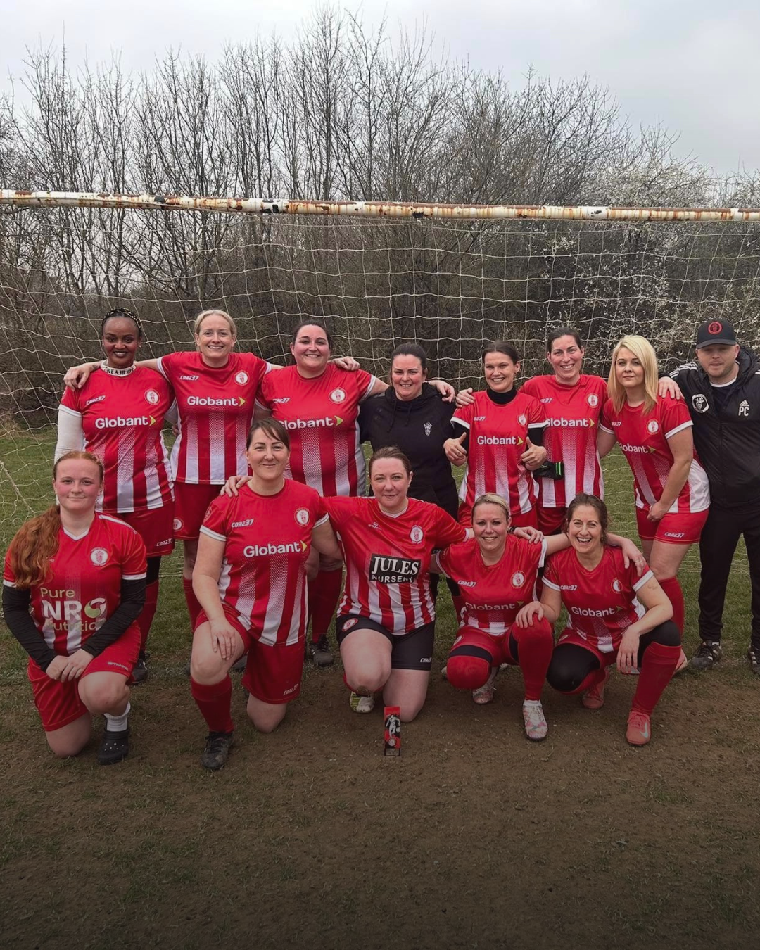 A women's soccer team posing in front of a goalpost on a field, wearing red and white striped jerseys.
