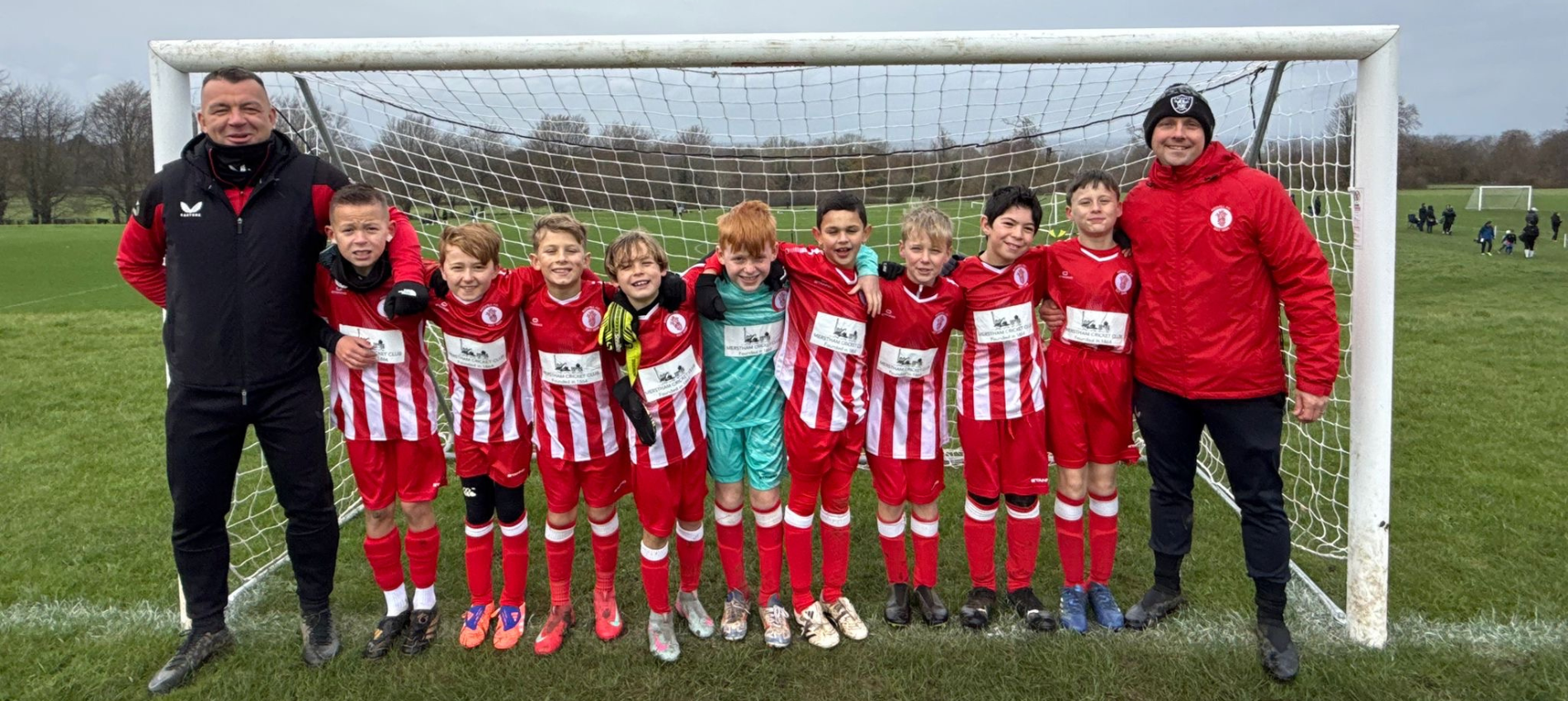 A youth football team with players and two coaches standing in front of a goal on a grassy field. The players are in red and white striped kit, and the coaches are wearing black and red jackets. The team looks happy and is posing for a group photo.