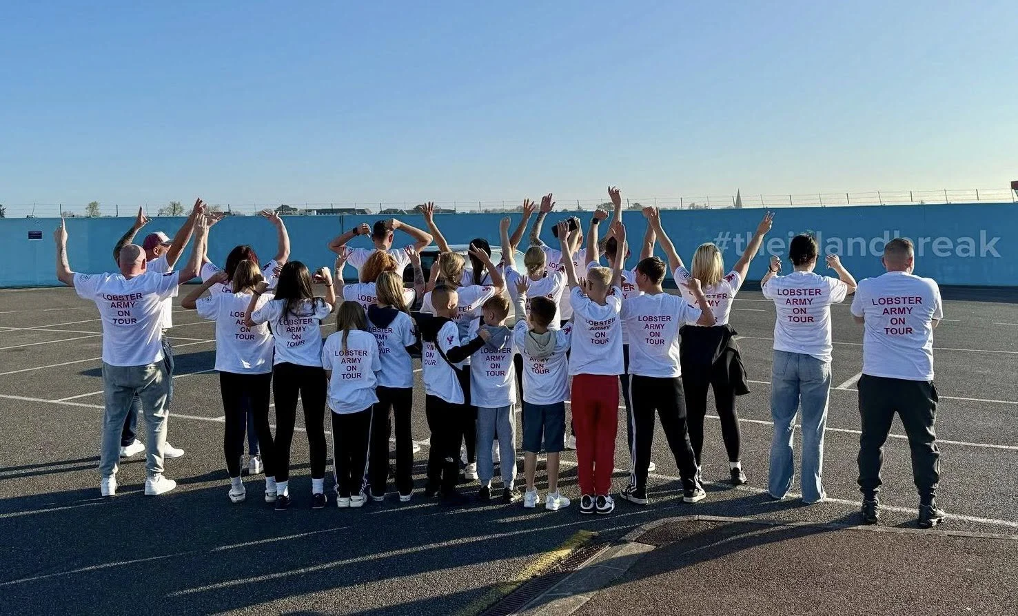Group of children and adults wearing white T-shirts with purple text, standing in a parking lot celebrating with raised hands during daytime.