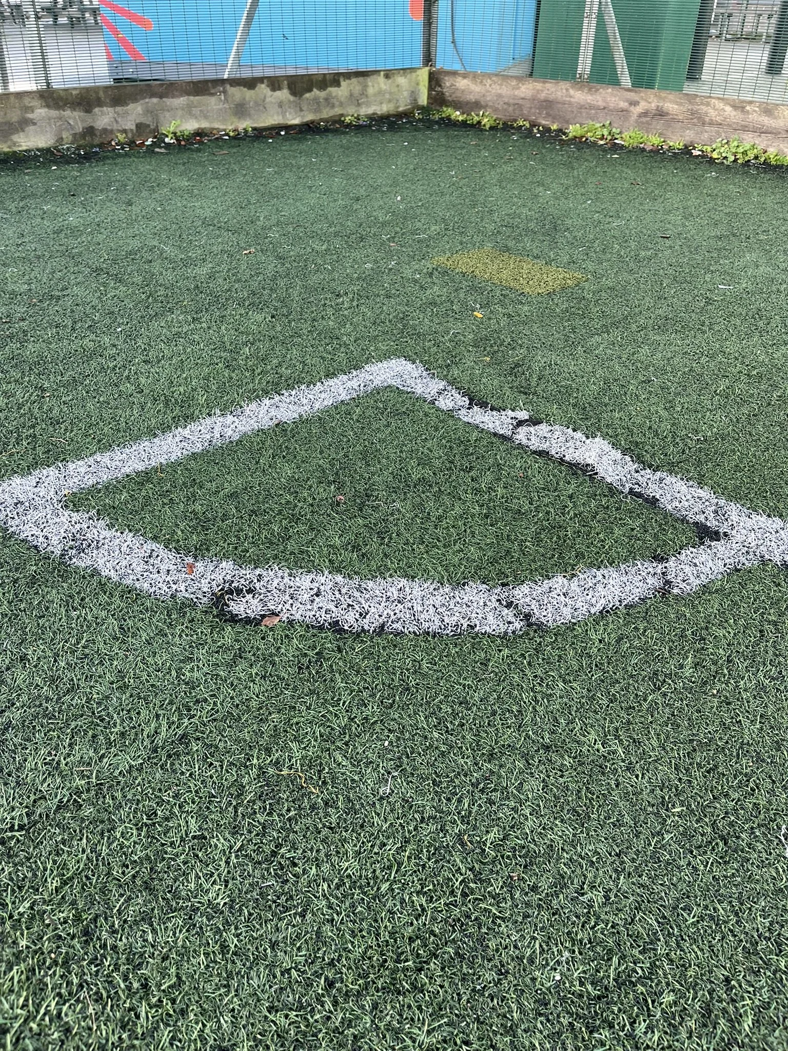 A small, rectangular soccer goal outlined with white paint on artificial turf at a sports field, with a yellow square in the background.