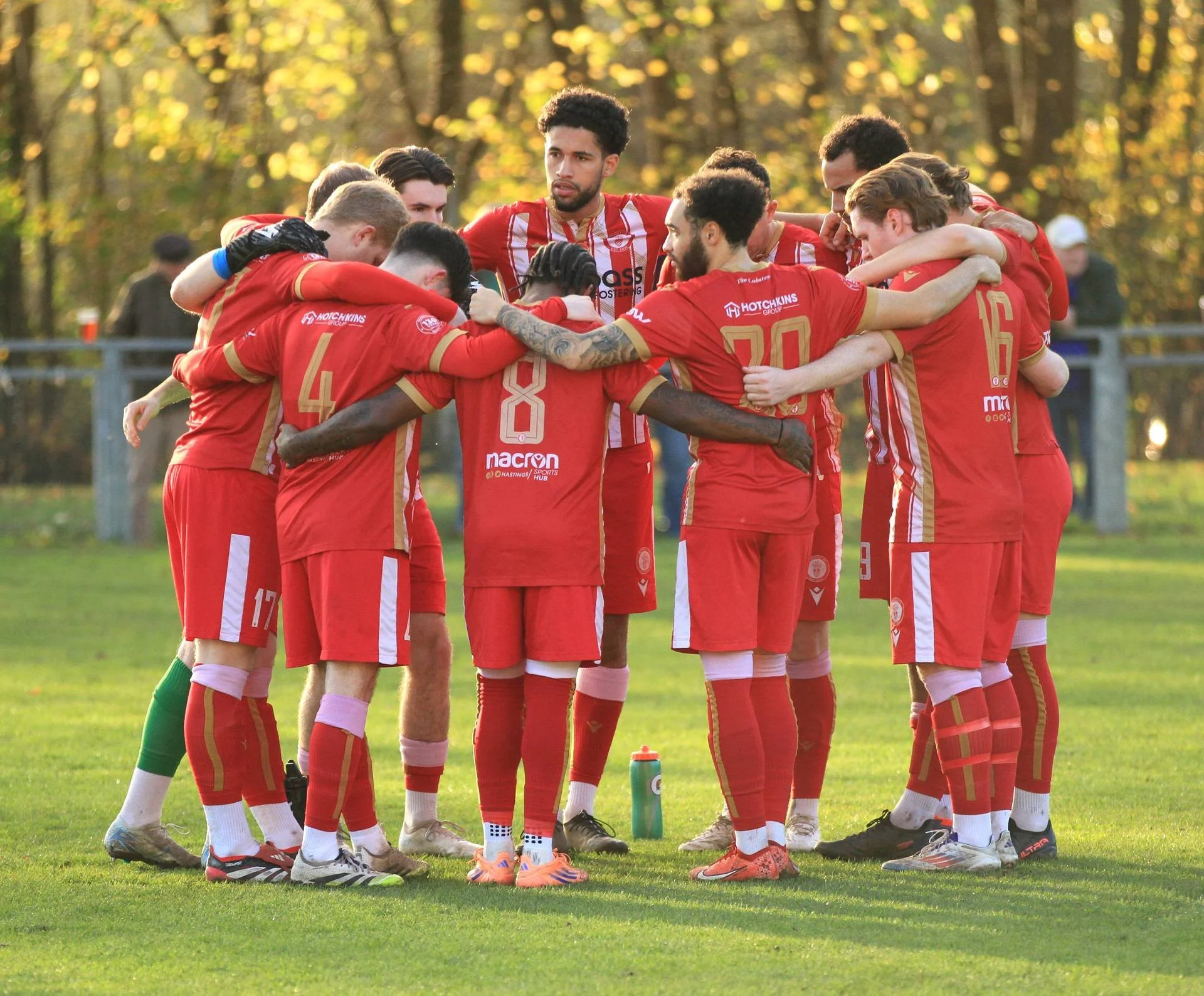 A group of Redhill FC football players in red kit huddled together on a pitch, likely in a team talk or pep talk before a game, with trees in the background and a water bottle on the ground.