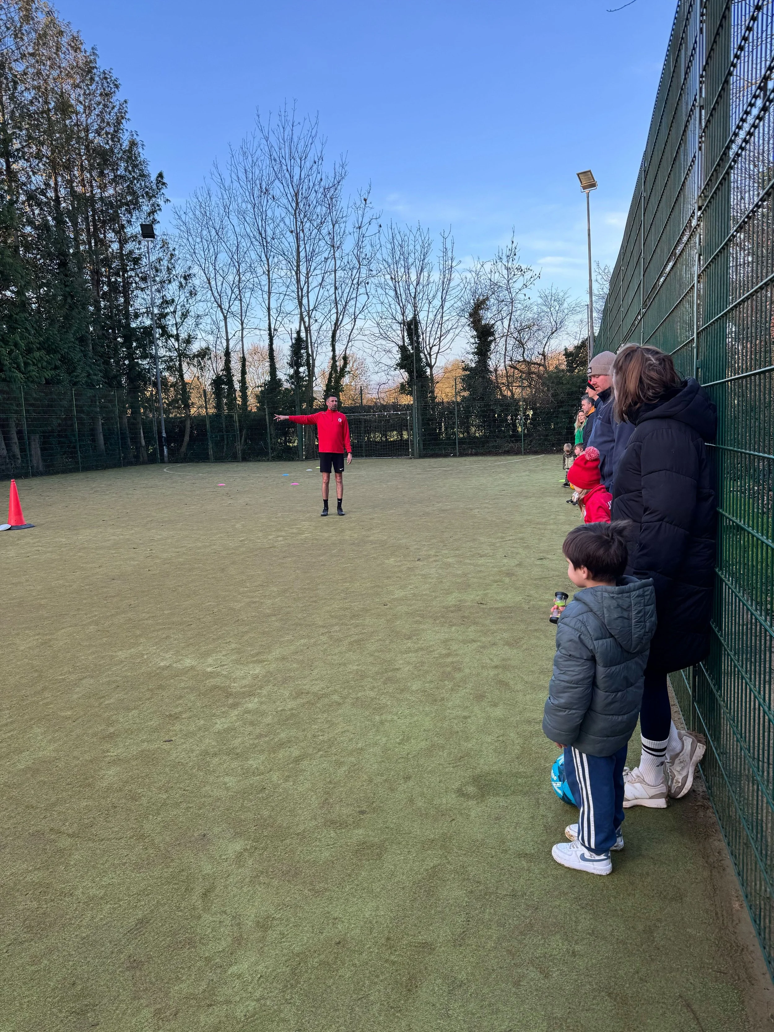 A soccer coach in a red shirt giving instructions to children during a training session on an outdoor field with a green fence and leafless trees in the background.