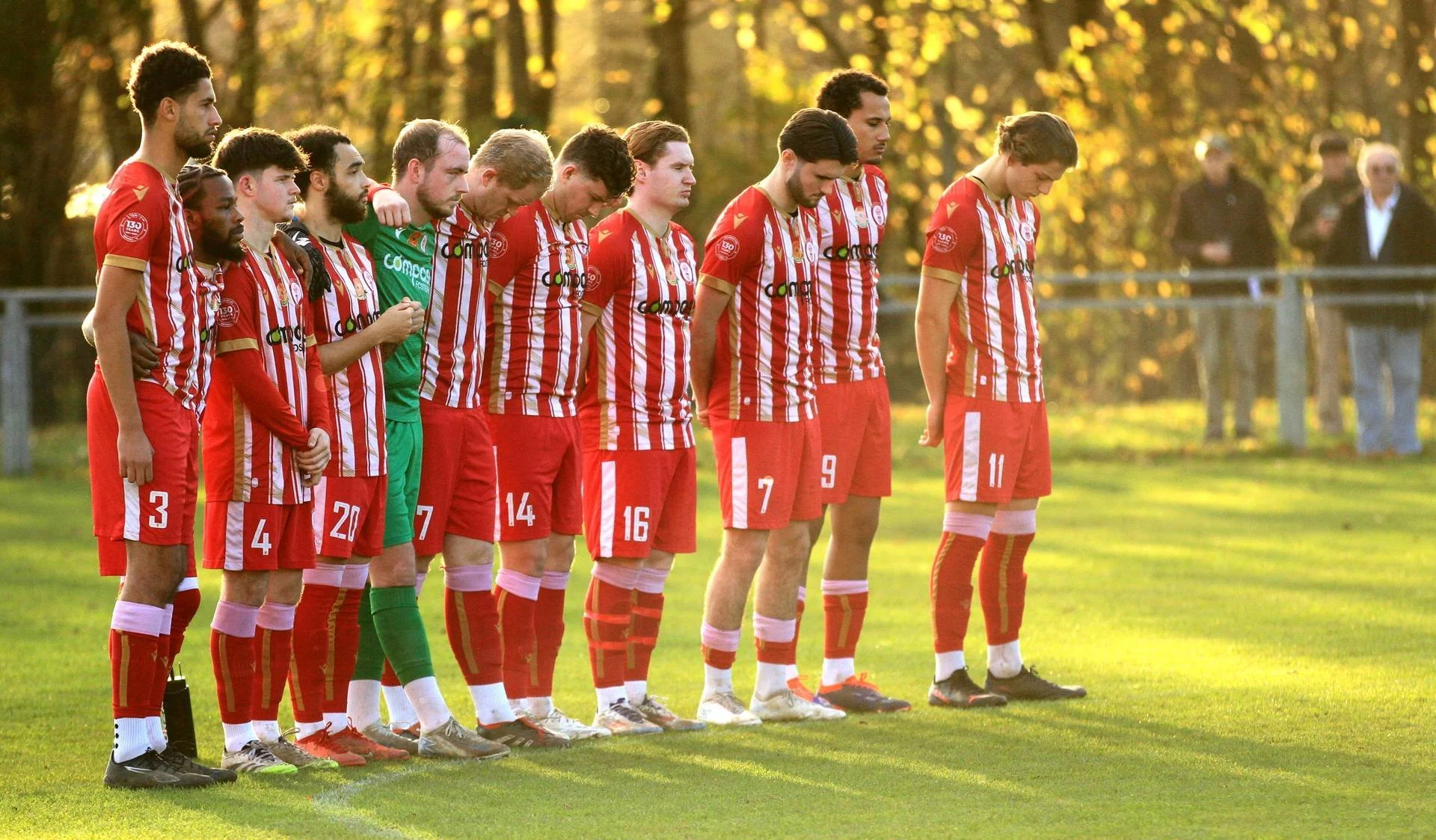 A soccer team in red and white striped uniforms standing in a line with heads bowed during a moment of silence on a grassy field in the late afternoon or early evening, with trees and spectators in the background.