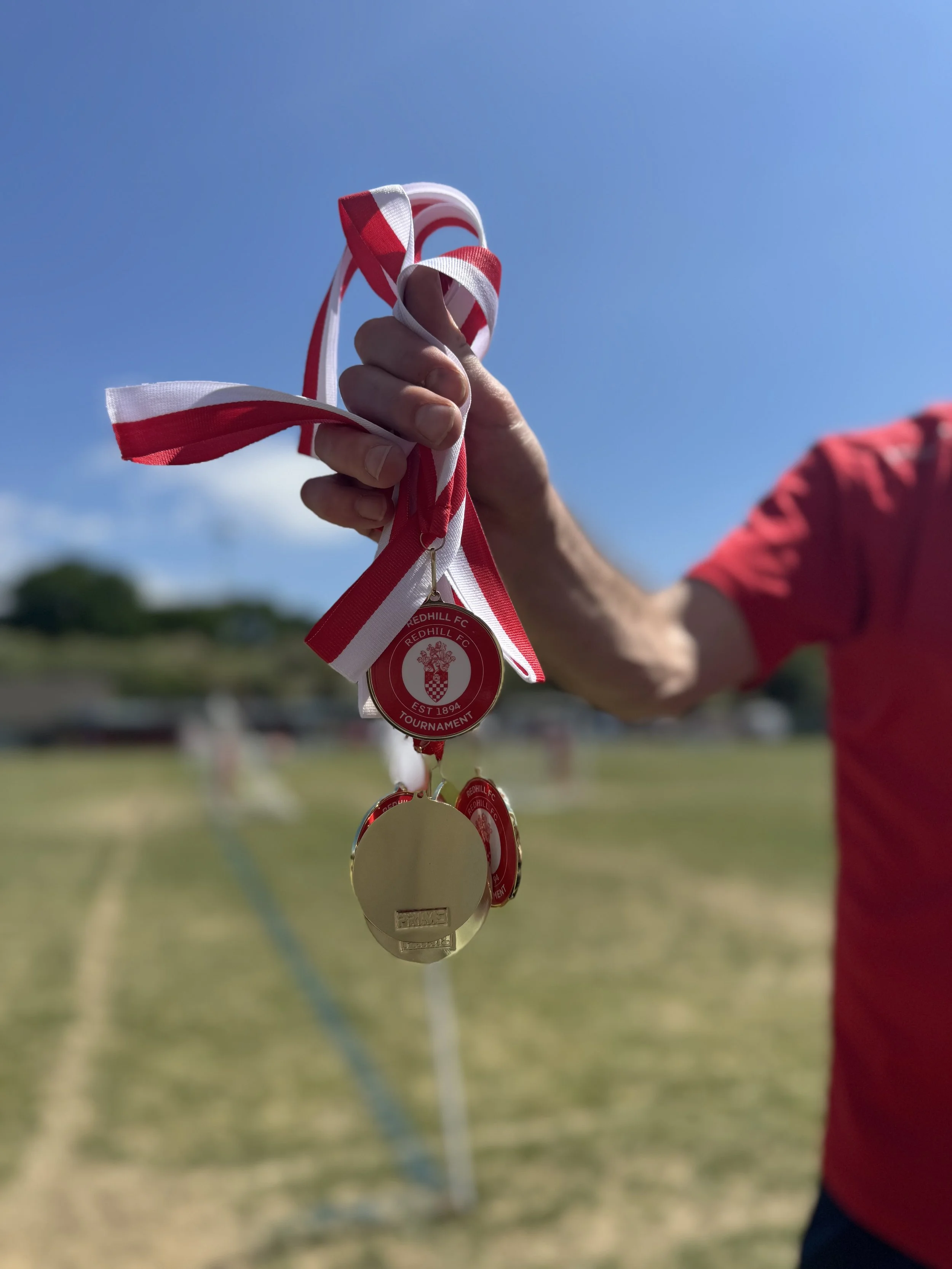A person holding a collection of medals on red, white, and striped ribbons, with a blue sky and a sports field in the background.
