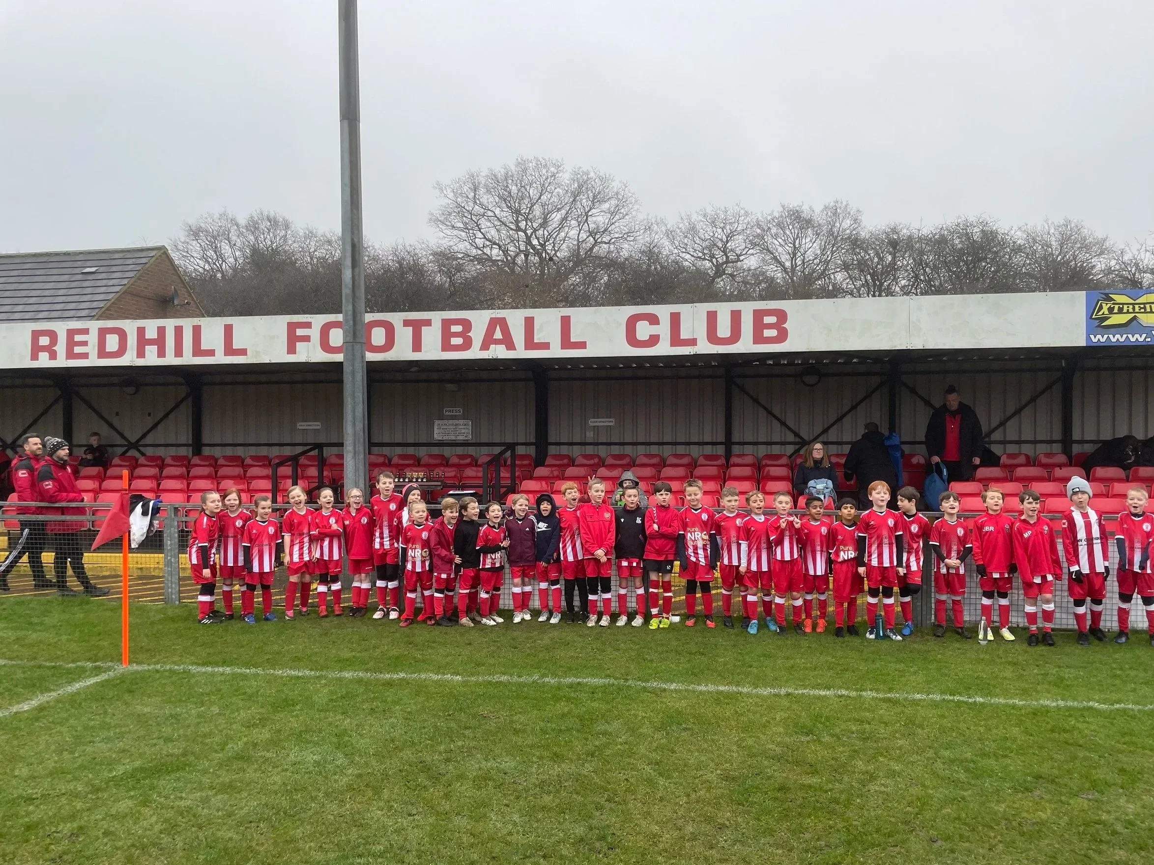 A group of young soccer players in red and white uniforms lined up on a soccer field, posing for a photo in front of the Redhill Football Club stand.