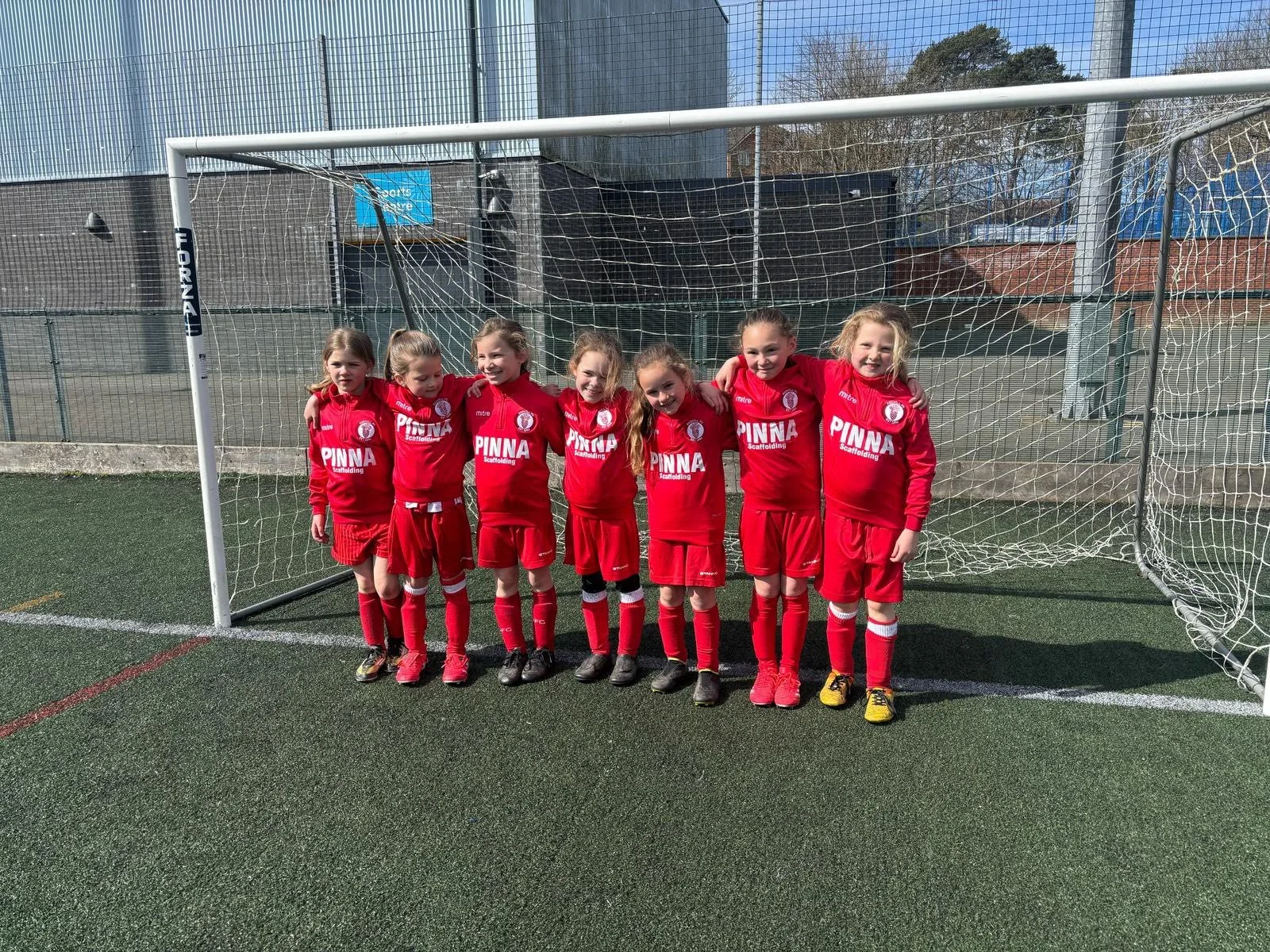 Group of seven young girls in red soccer uniforms standing together in front of a goalpost on a soccer field, smiling with their arms around each other.