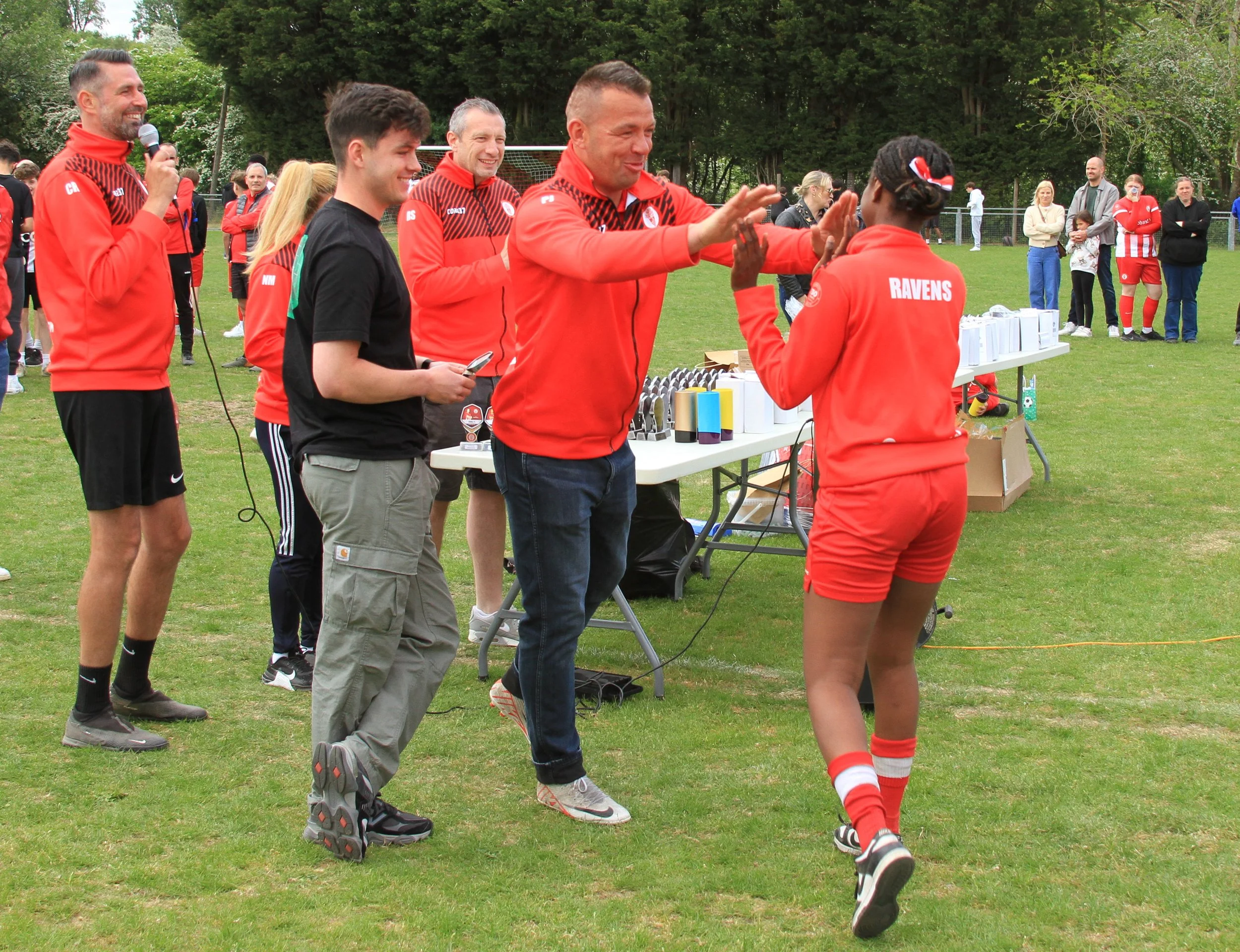 A girl in a red sports kit receives a high-five from a man with a red jacket, with other people in red sportswear and casual clothes in the background at an outdoor event on a grassy field.