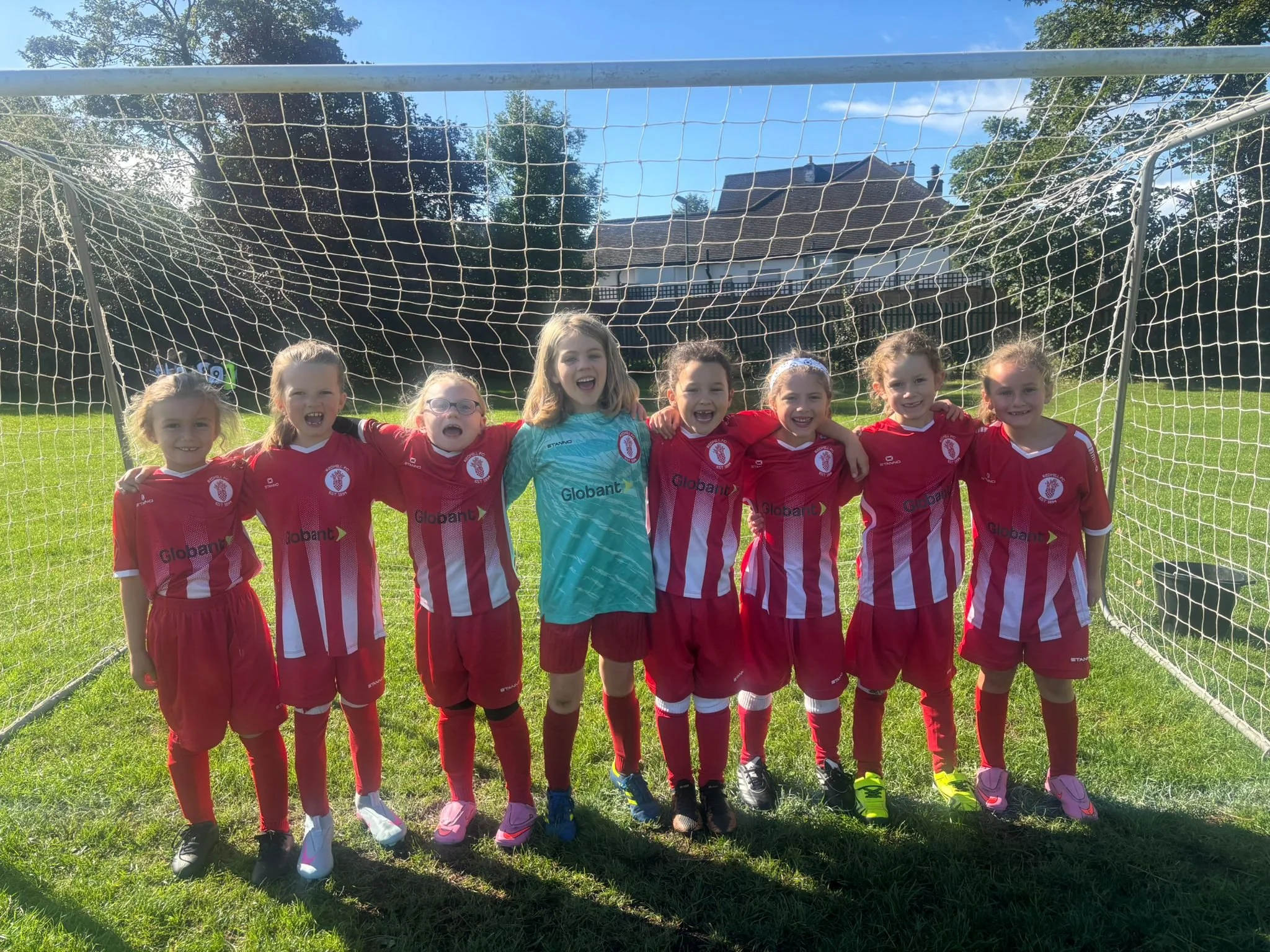 Children in red soccer uniforms standing in front of a soccer goal on a grassy field, smiling for a group photo.