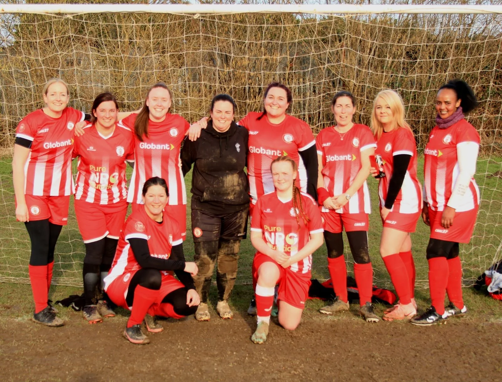 A group of women football players in red and white kits, posing on a football pitch with a goalpost behind them. They are smiling and standing on grass with a wooded area in the background.