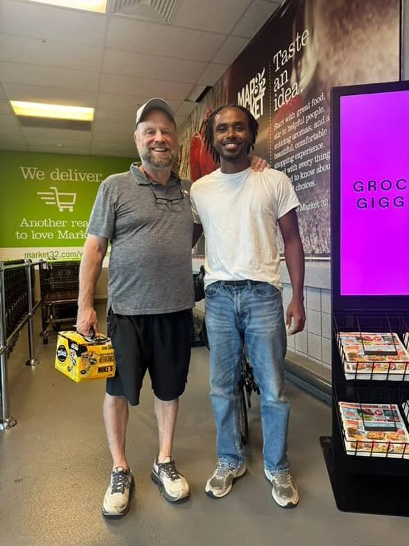 Two men standing together inside a grocery store, smiling at the camera. One is holding a yellow box of snacks. There are grocery carts and store signs in the background.