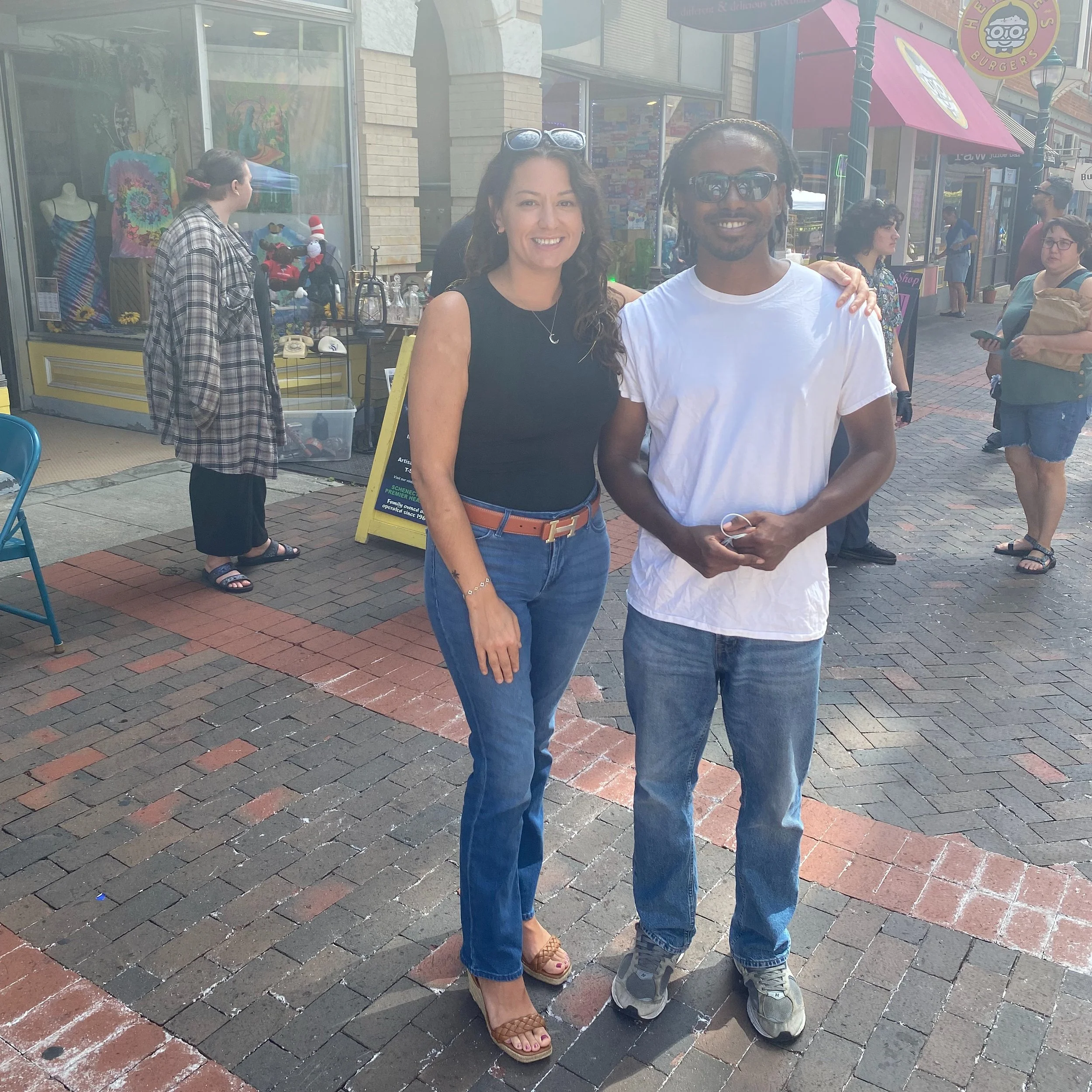 A woman in a black sleeveless top and blue jeans standing next to a man in a white t-shirt and blue jeans on a brick sidewalk outside a store. Several people are in the background.
