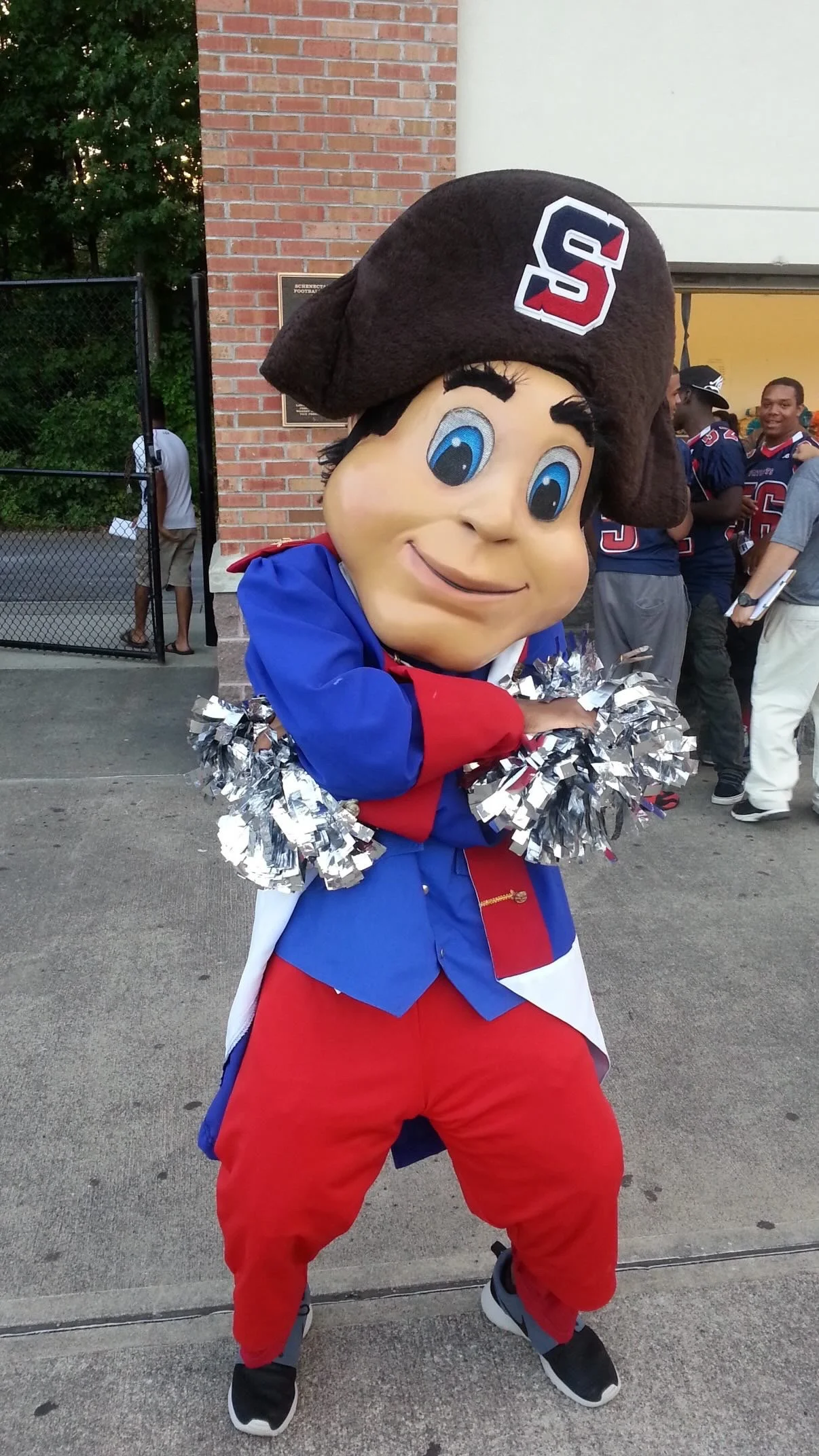 Person in a mascot costume dressed as a cheerleader with pom-poms, wearing a pirate-themed costume with a large head, big blue eyes, and a brown pirate hat with a sports team logo.