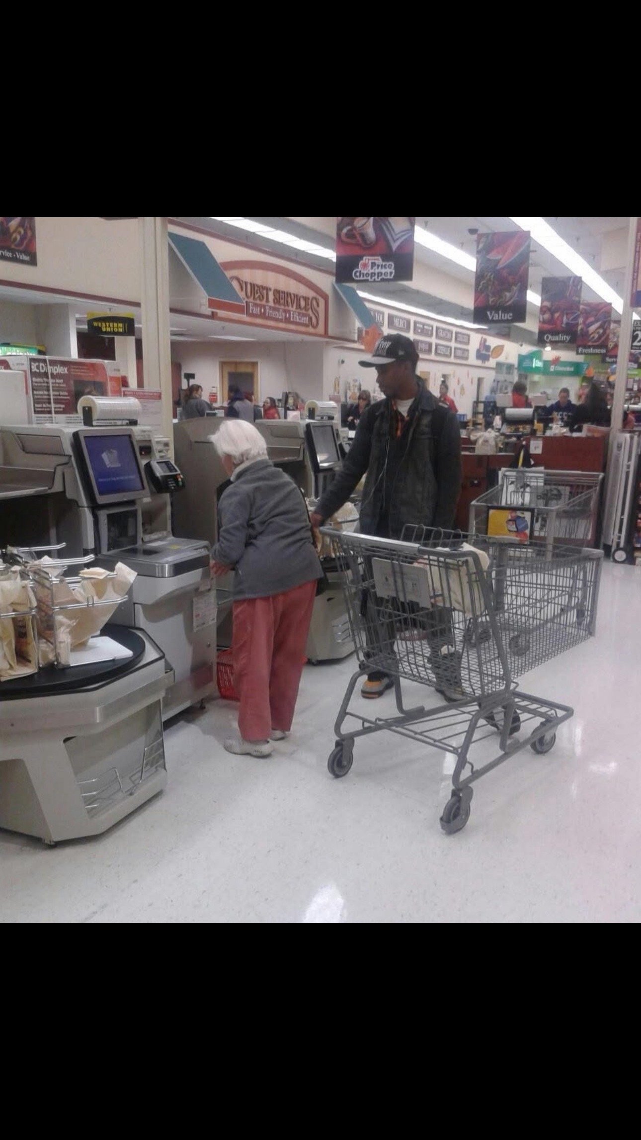 An elderly woman and a young man are shopping at a grocery store checkout lane. The elderly woman is using a self-checkout machine, and the young man is standing behind her, holding a shopping cart. The store is well-lit, with various signs and other