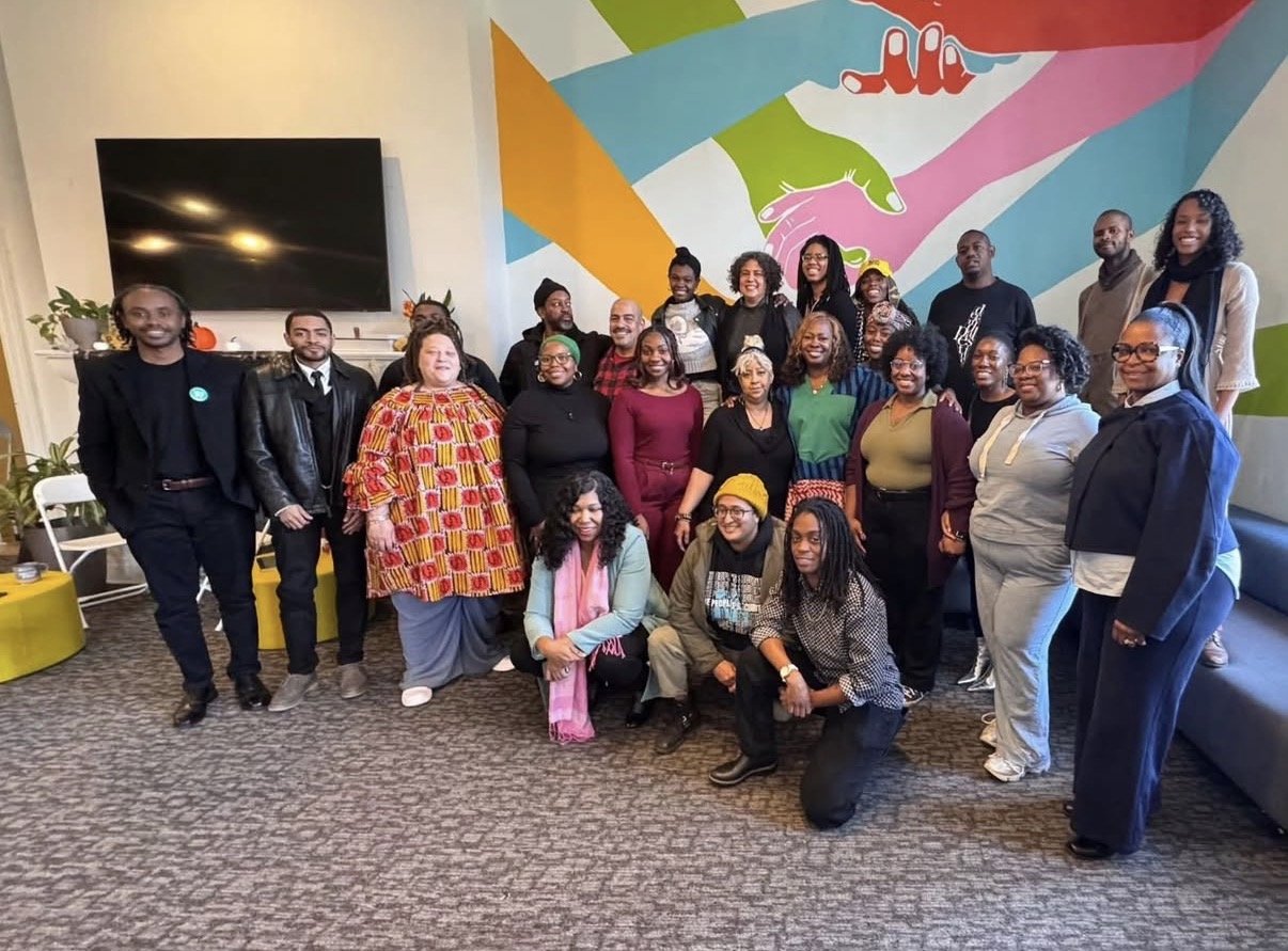 Group of people posing together in an indoor room with a colorful mural on the wall behind them.