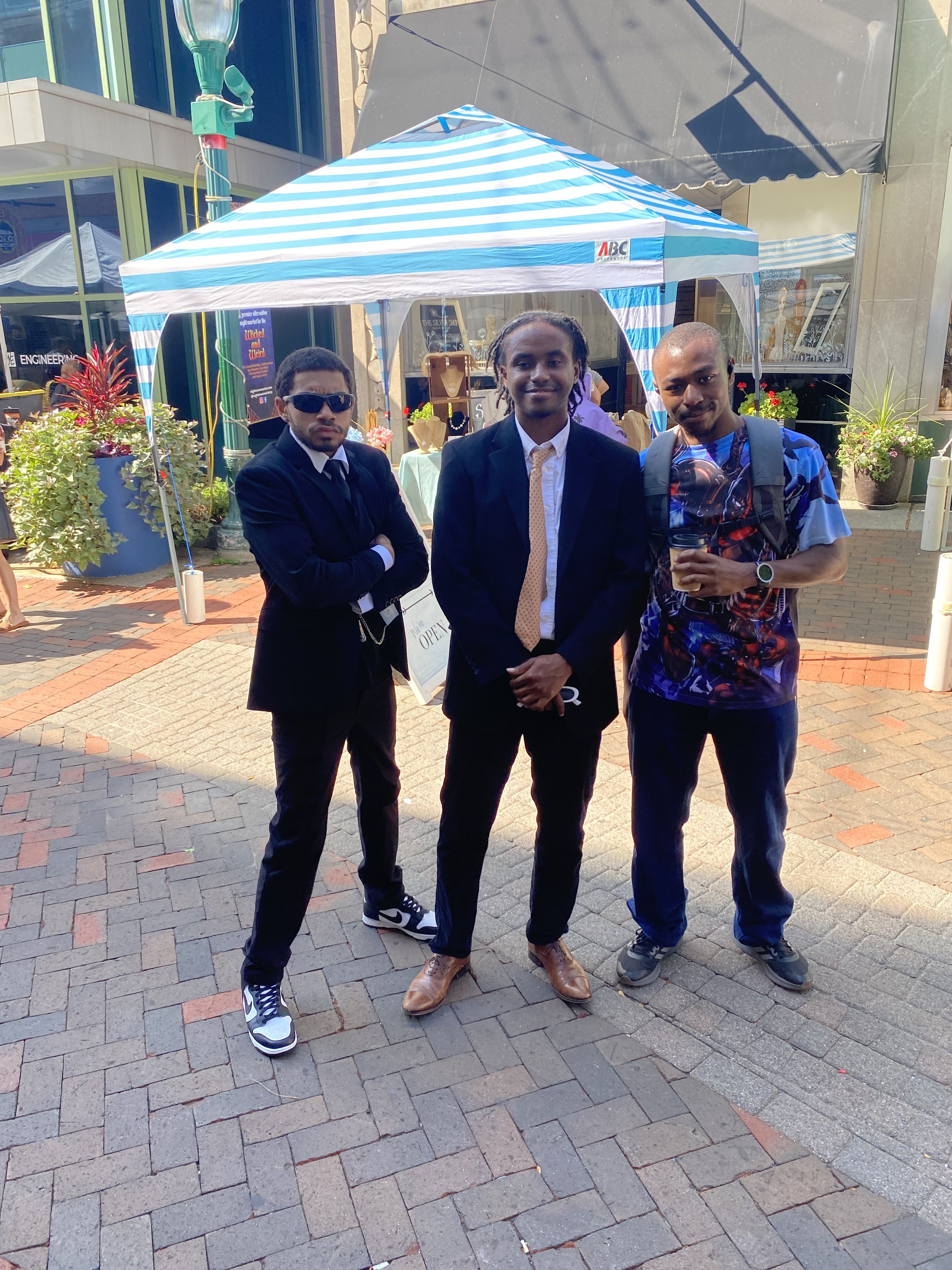 Three men standing in front of a booth with a striped canopy at an outdoor event in an urban setting.