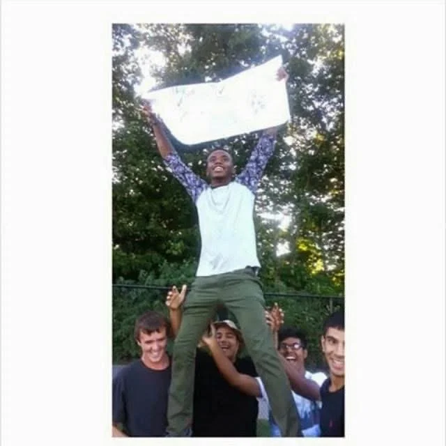 A young man standing on the shoulders of four friends, holding a large white board or sign above his head, outdoors among trees.