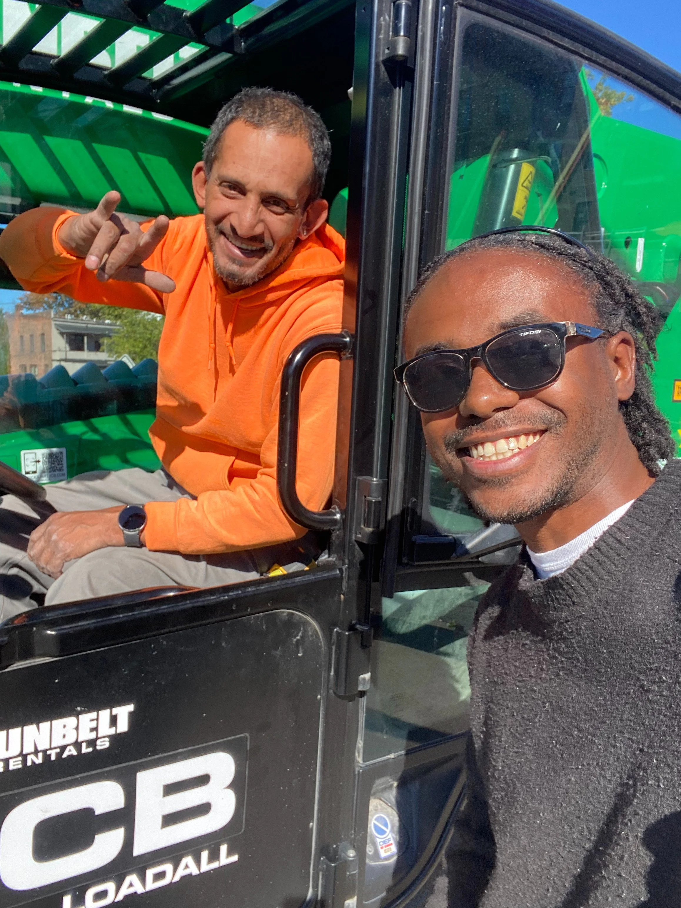 Two smiling men, one sitting in a green utility vehicle and the other standing outside, posing for a selfie on a bright, sunny day.