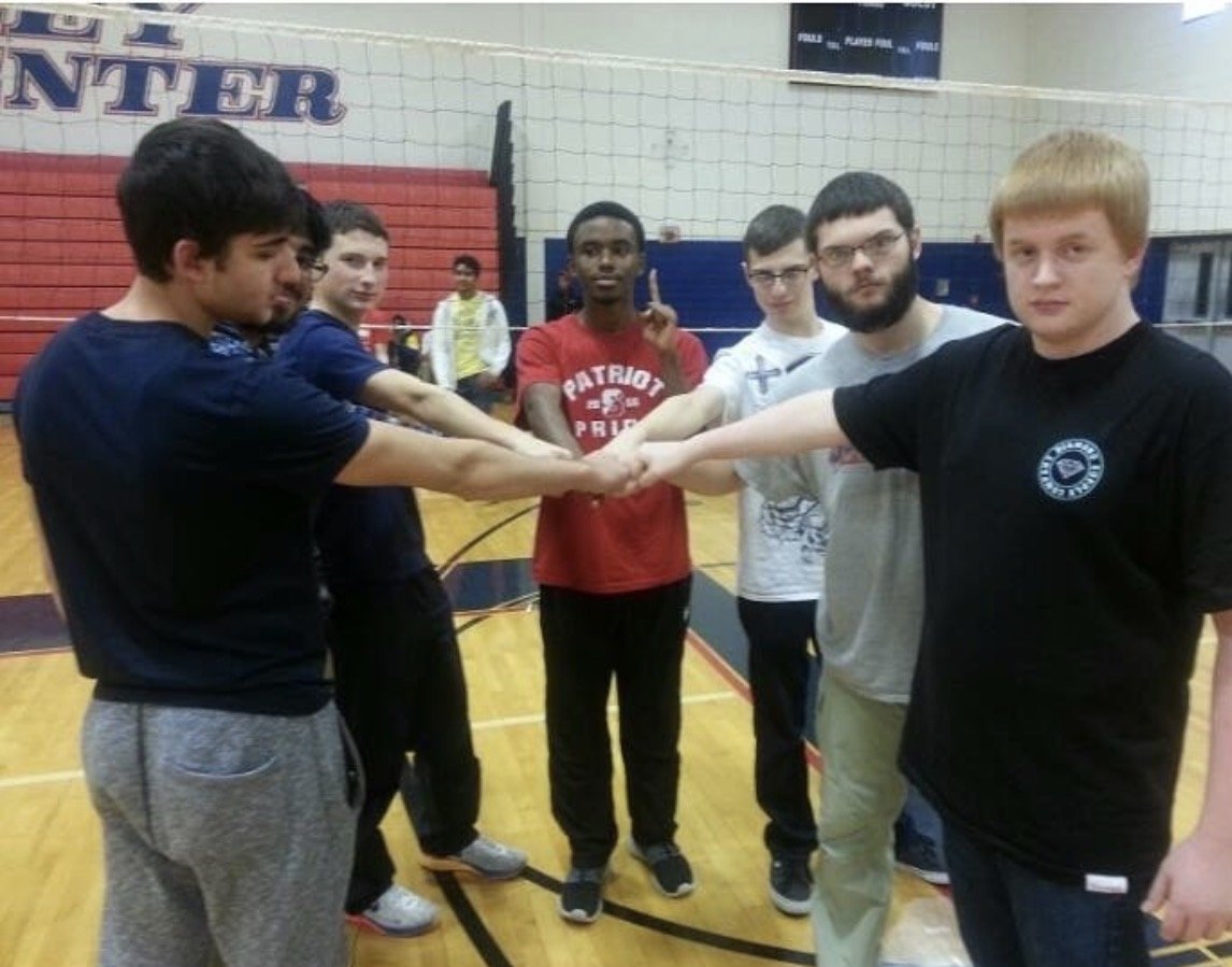 Six young men standing in a gymnasium on a volleyball court, with their hands stacked together in the center, expressing teamwork and unity.