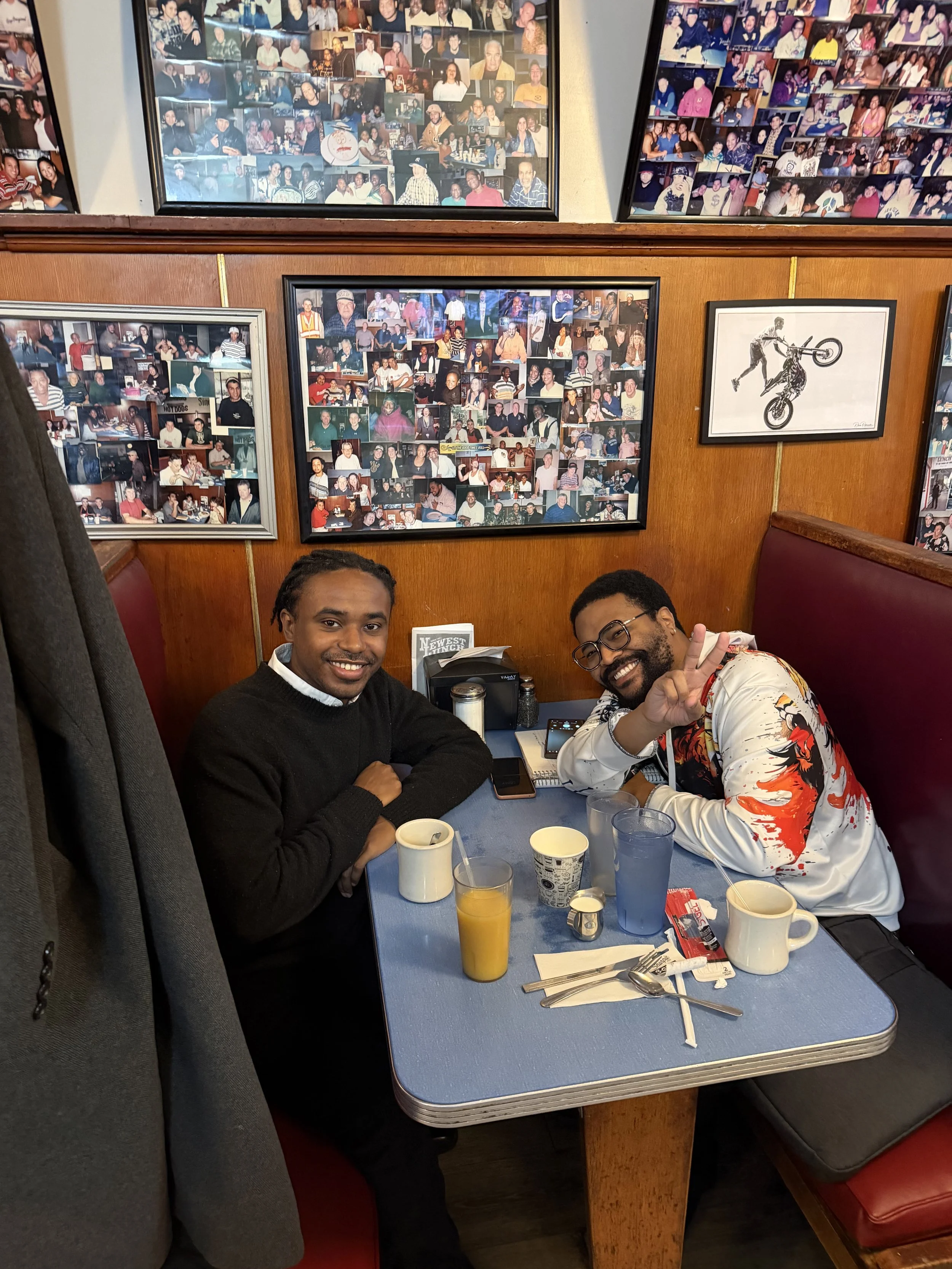 Two men sitting at a restaurant table, smiling at the camera. One is wearing a black sweater, and the other is wearing a white jacket with red and black design. The table has drinks, utensils, and cups, with photographs on the wood-paneled wall behin