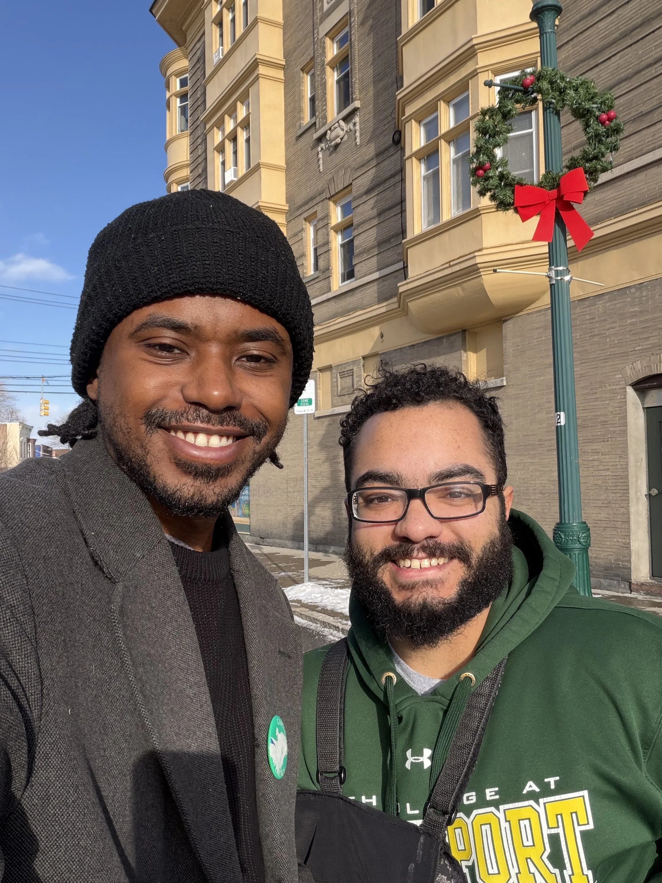 Two men are outdoors taking a selfie in front of a yellow brick building decorated with a Christmas wreath with red bow and berries on a green street lamp. The man on the left wears a black beanie and gray coat, and the man on the right wears glasses