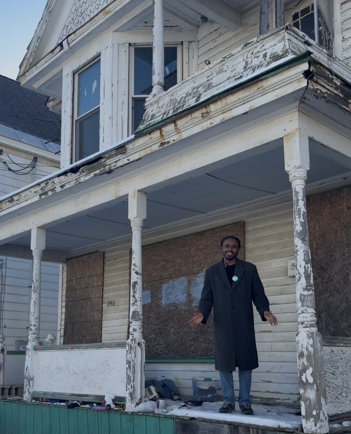 A man standing on the porch of a dilapidated house with peeling paint. The house has boarded-up windows and a second-story porch with a bay window. The man is smiling and gesturing with his hands.