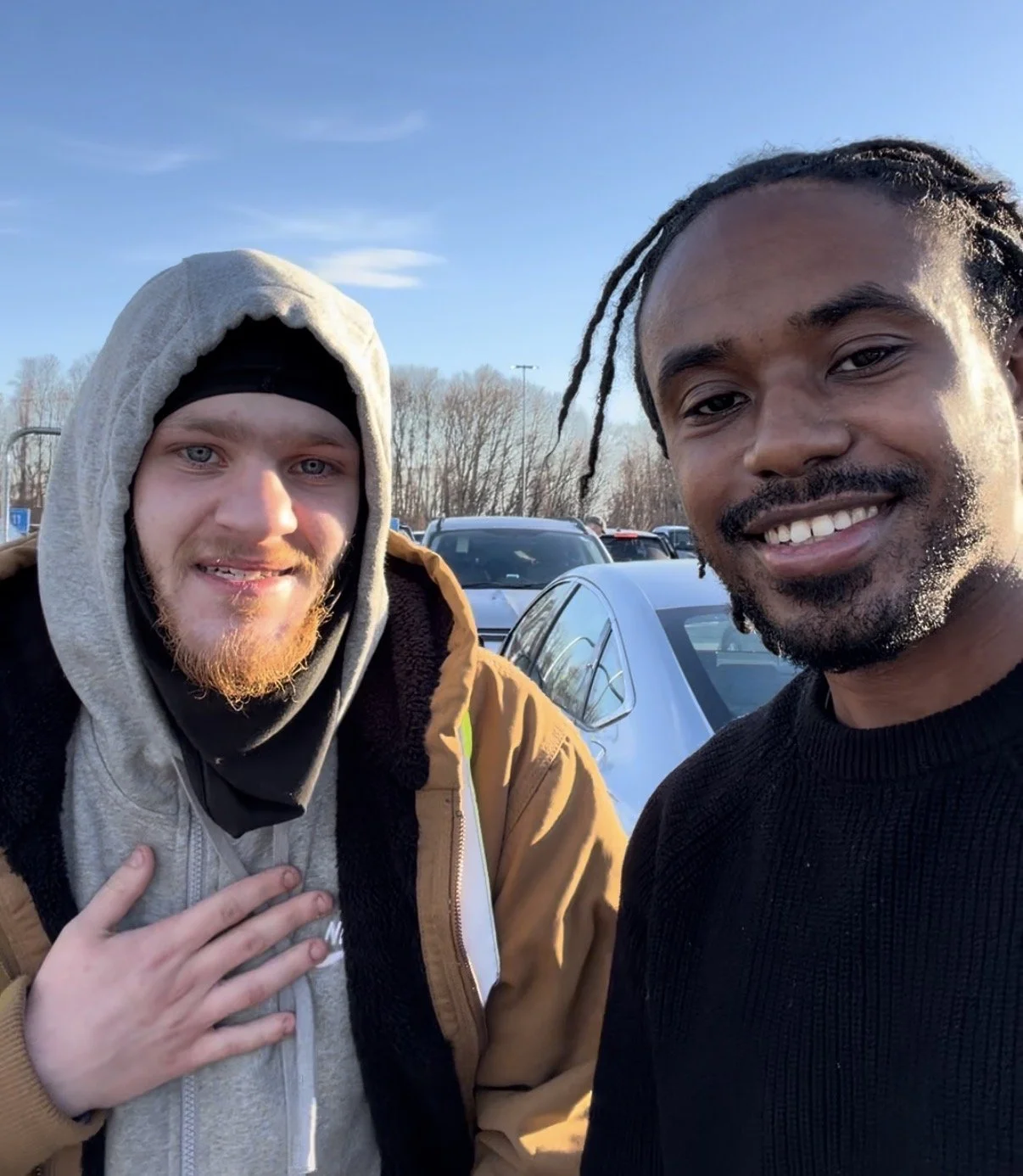 Two men taking a selfie in a parking lot on a sunny day. One man has a light beard, red hair, and is wearing a hoodie and a tan jacket. The other man has dark hair in braids and is wearing a black sweater. There are cars and trees in the background.