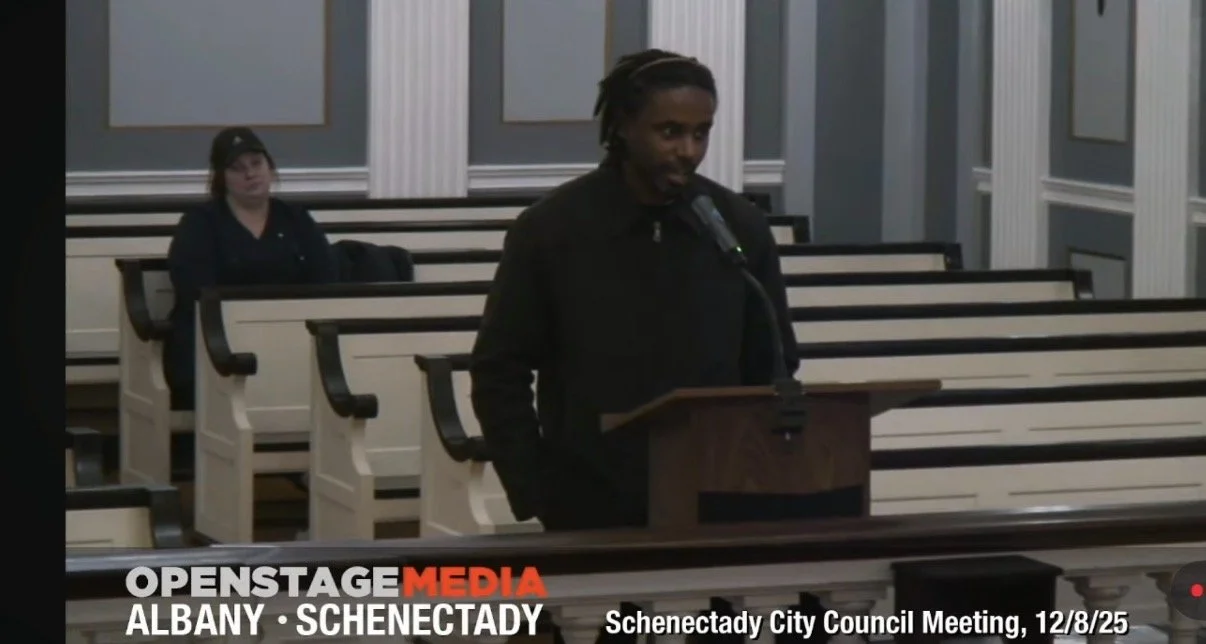 A man with dreadlocks speaking at a podium in a city council chamber with empty benches behind him, during the Schenectady City Council Meeting on December 8, 2025.