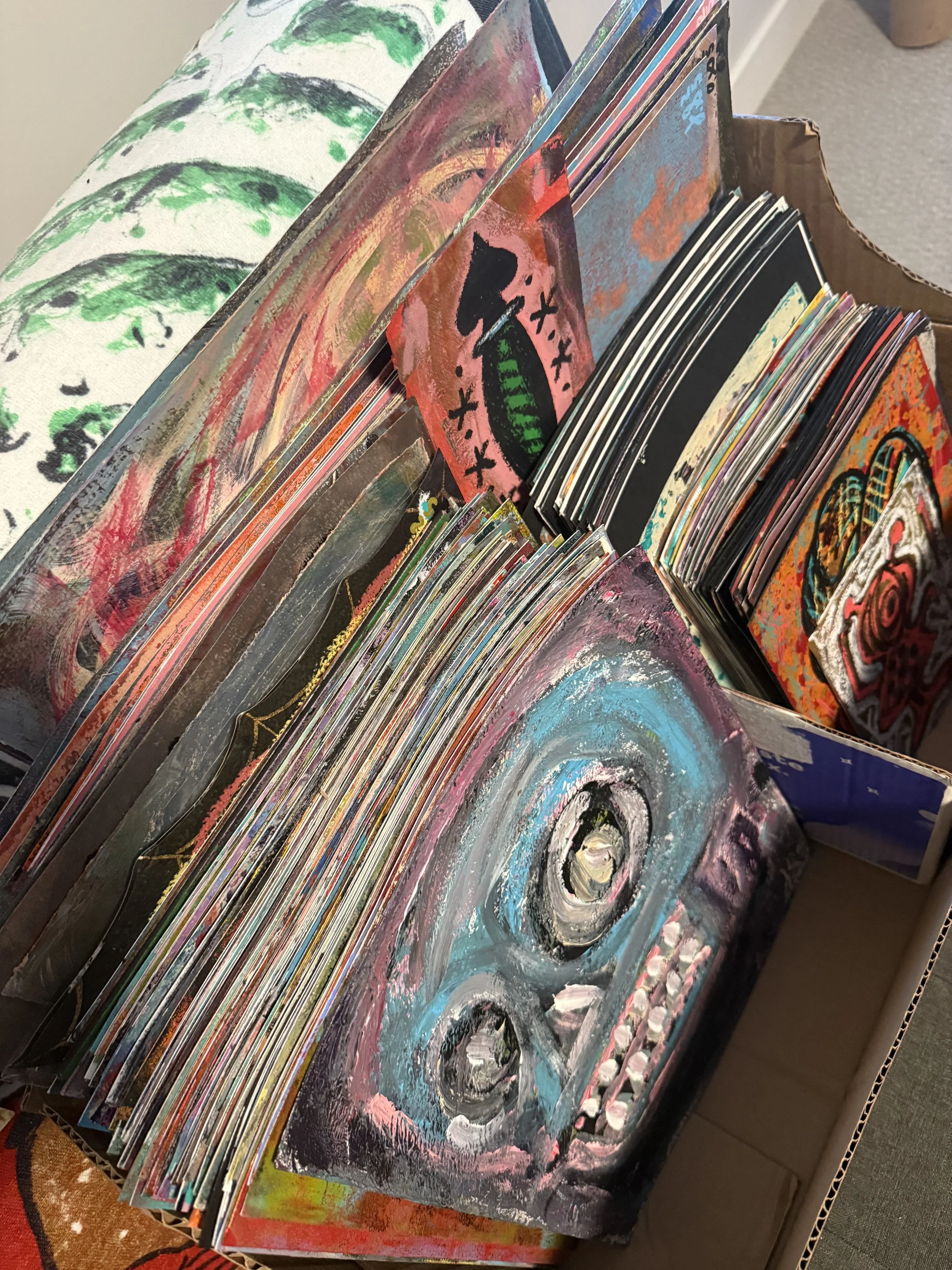 A cardboard box filled with colorful, abstract paintings and artwork, stacked together.